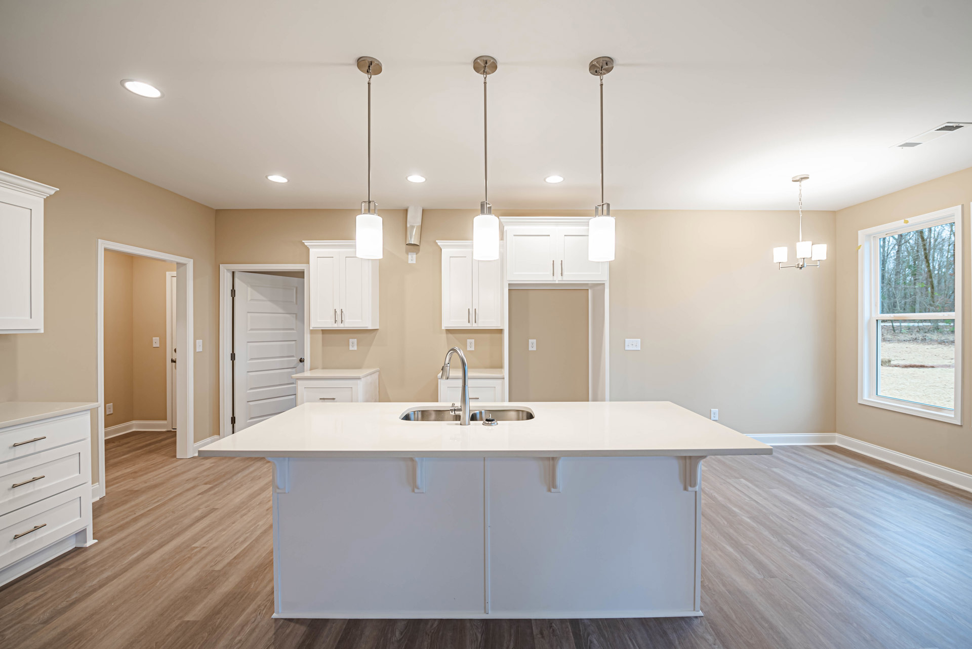 White kitchen island with built-in sink, wood flooring, white cabinetry with metal handles, pendant lights overhead, large window revealing trees outside