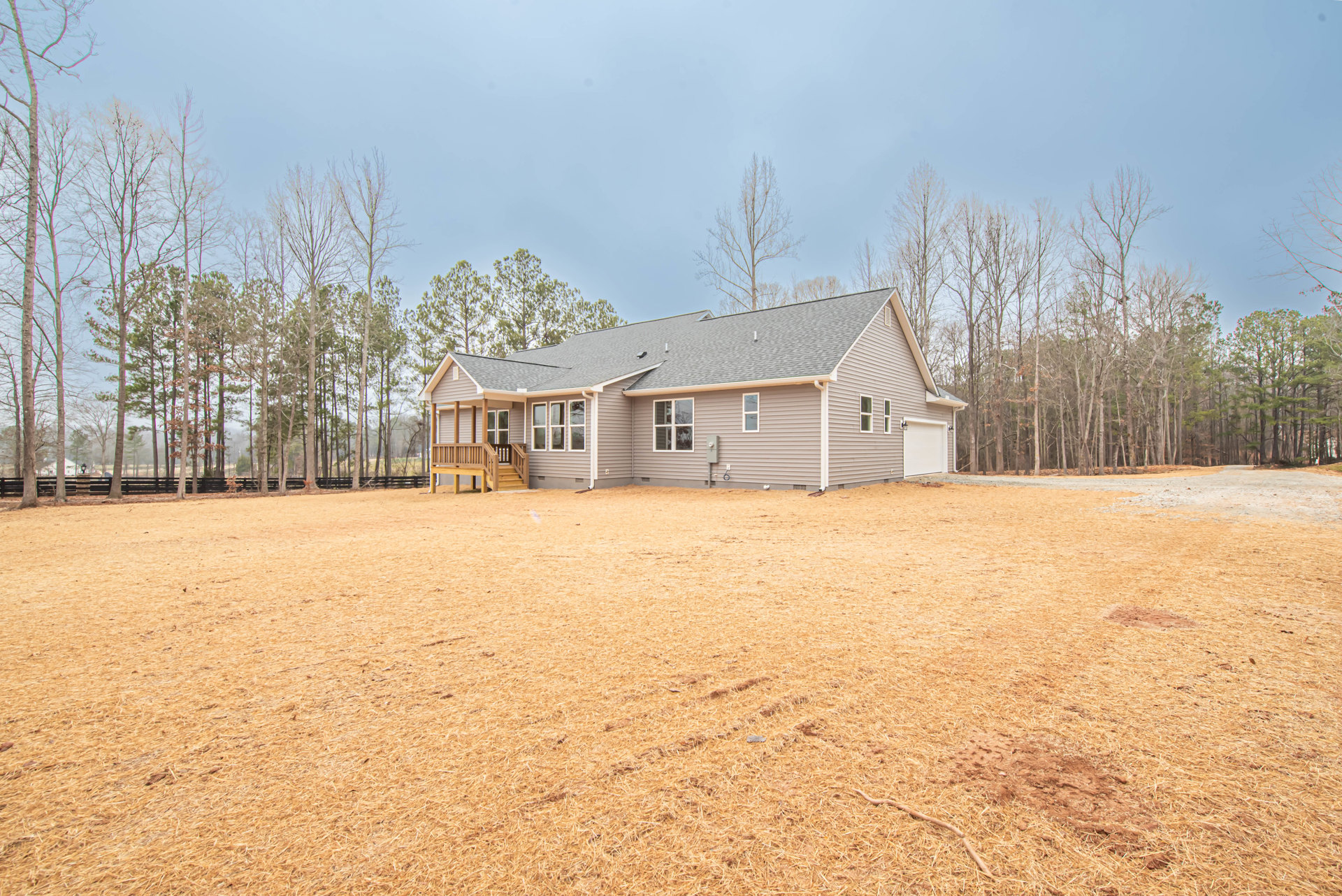 Two-story house with white-framed windows, wooden deck with railing, spacious grassy yard, mature trees in background, partly cloudy sky