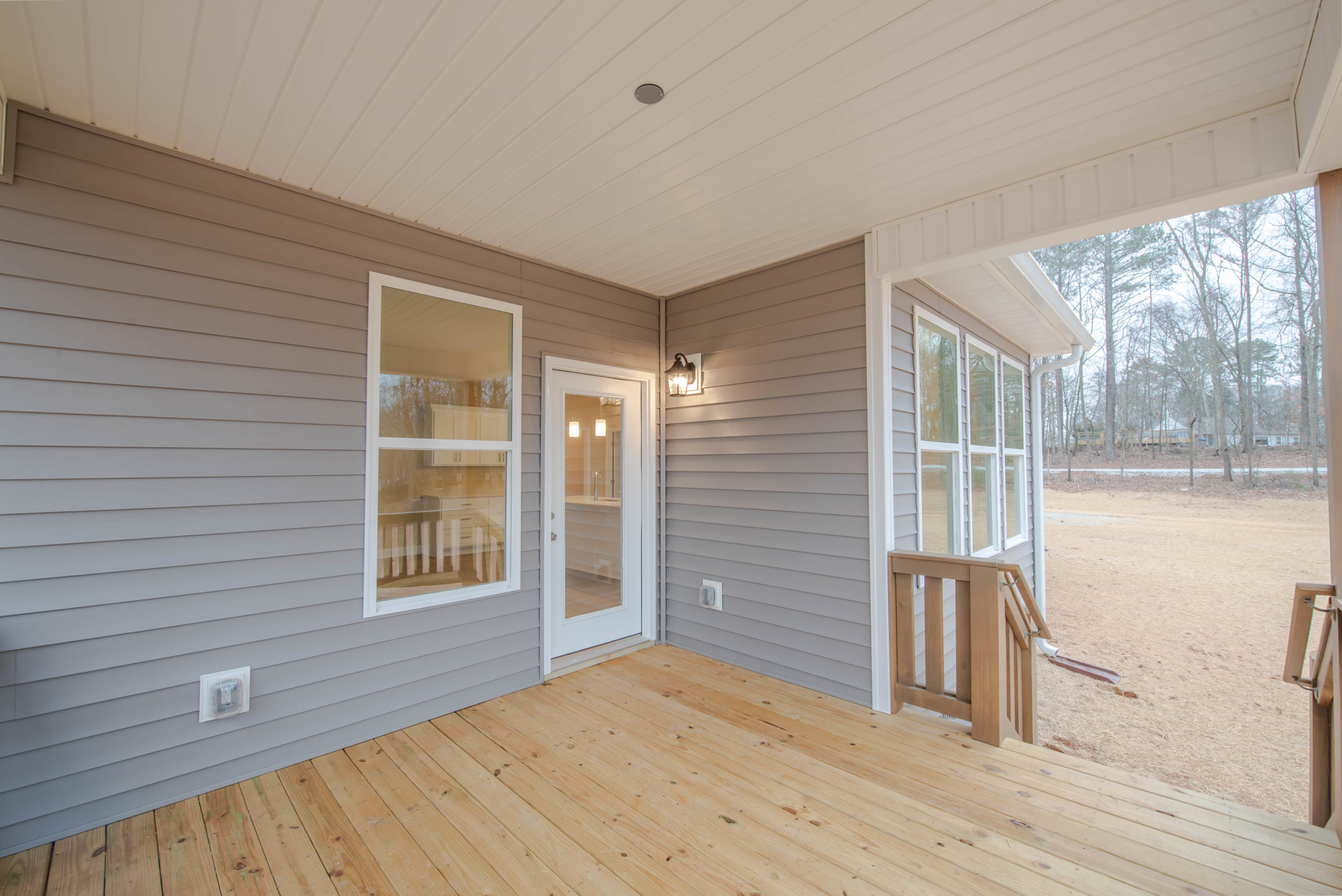Wood deck with plank flooring, glass-paneled door, grey siding exterior, porch area, ceiling light fixture, and large window
