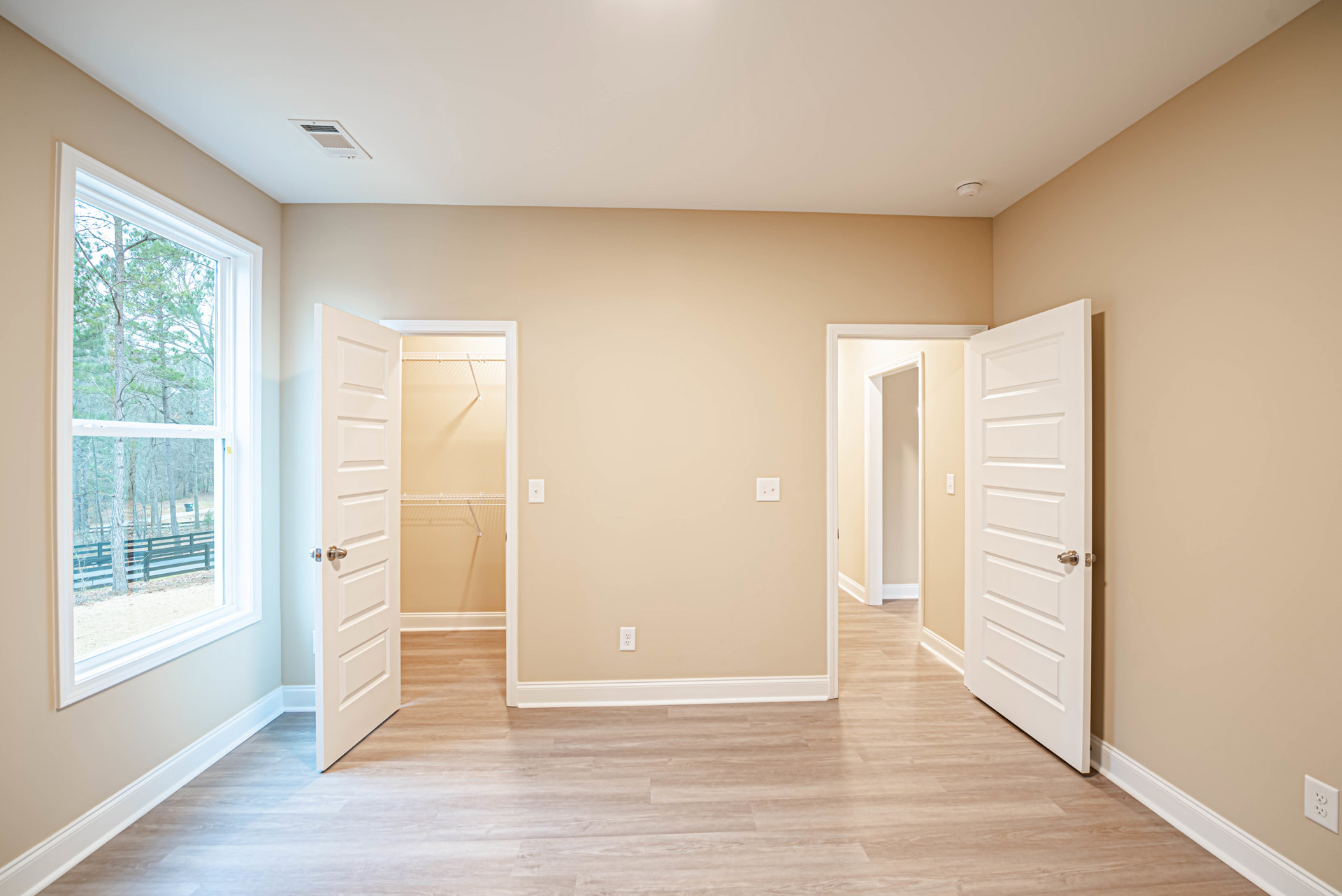 Open doorway leading to a room with light wood flooring, white walls, ceiling vent, and closet with white shelving; window reveals green trees outside.