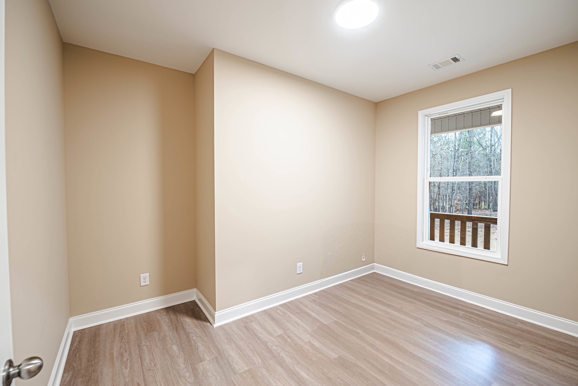 Sunlit room featuring a large window overlooking trees, hardwood flooring with white baseboards, plaster walls, and a close-up of a wooden railing and brushed metal doorknob.