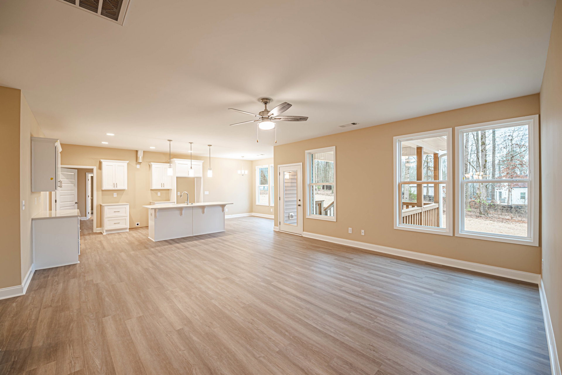 Spacious room with wood flooring, ceiling fan, white cabinets, glass door, large window overlooking trees, and a sink with white countertop