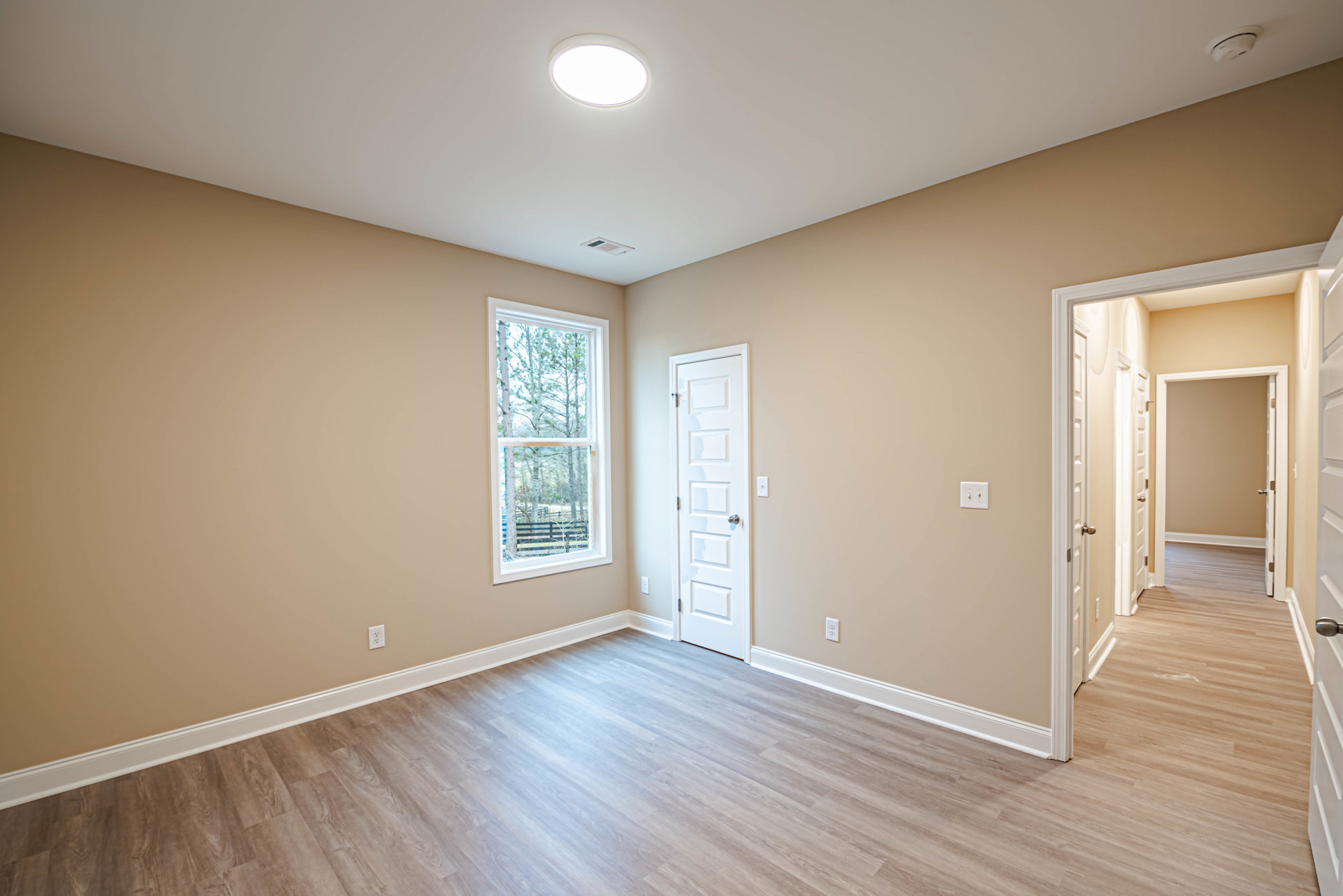 Bright room with wood flooring, white walls, white door with silver doorknob, large window showing trees outside, circular ceiling light fixture, white baseboard molding