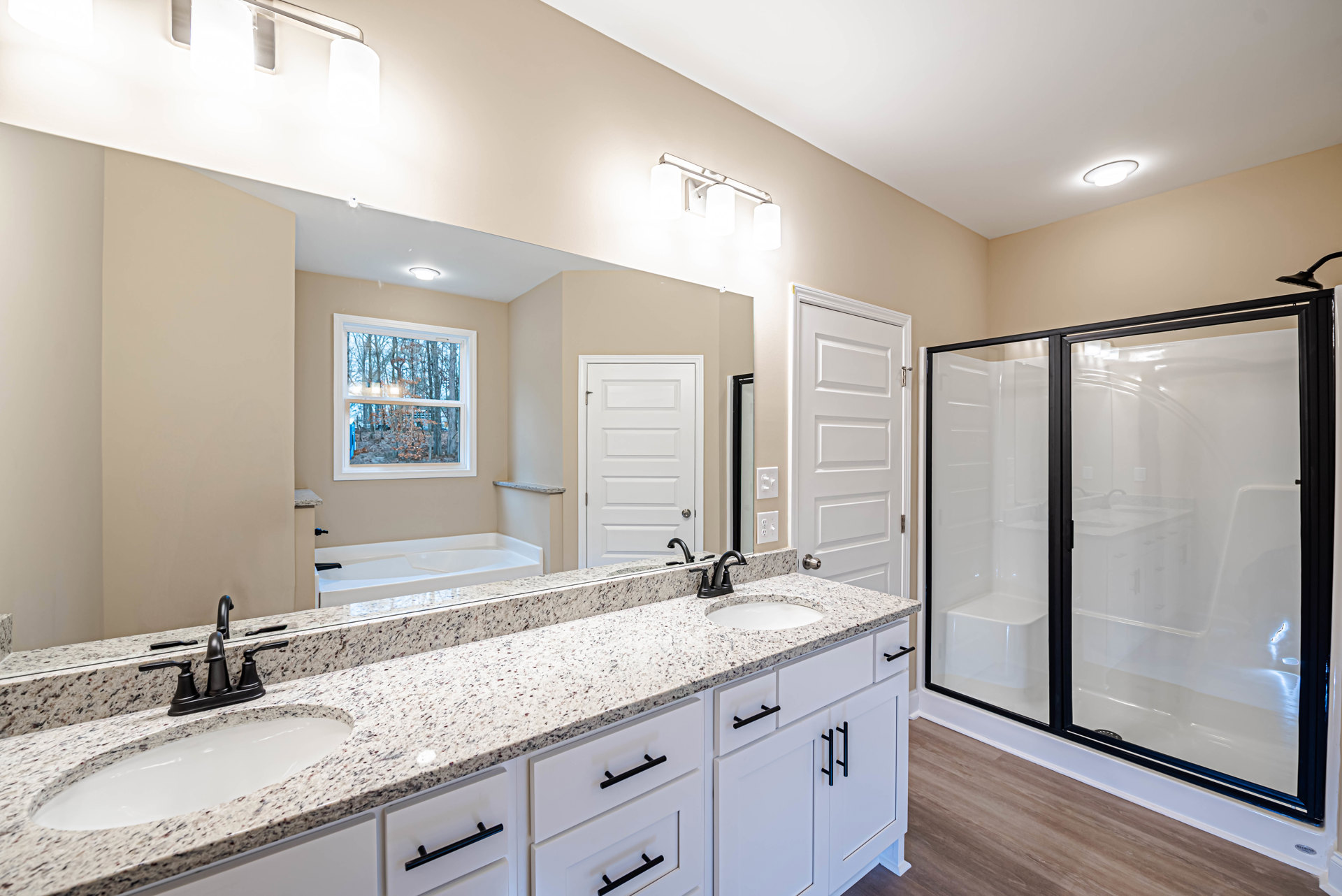 Bathroom with glass-enclosed shower, double sink vanity with stone countertop and drawers, large mirror, white door with silver handle, window overlooking trees.