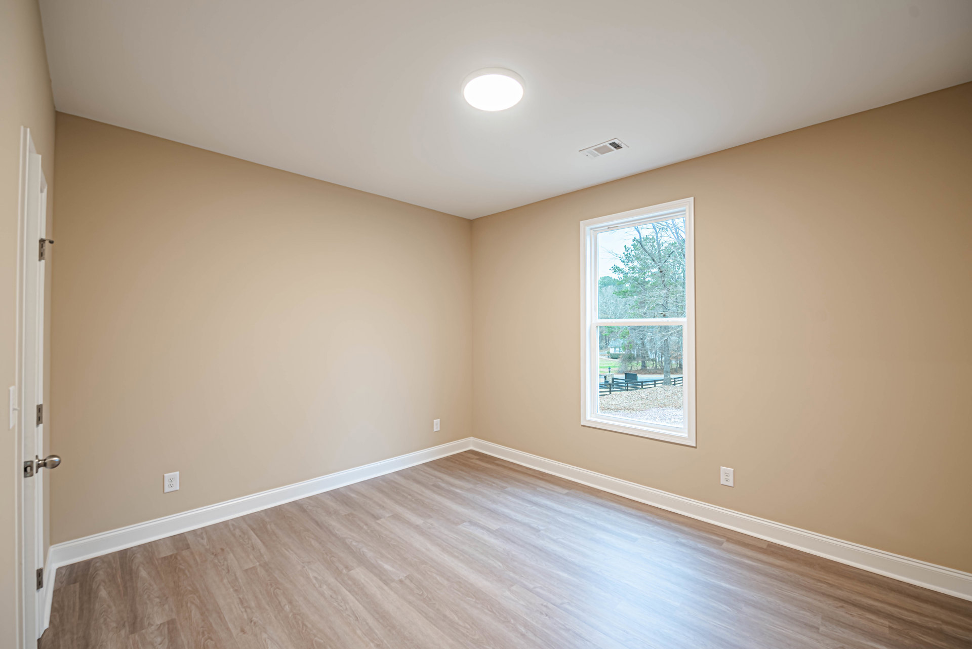 Sunlit room featuring wide-plank hardwood flooring, large window overlooking leafy trees, smooth plaster walls, white ceiling with recessed light fixture, and simple baseboard