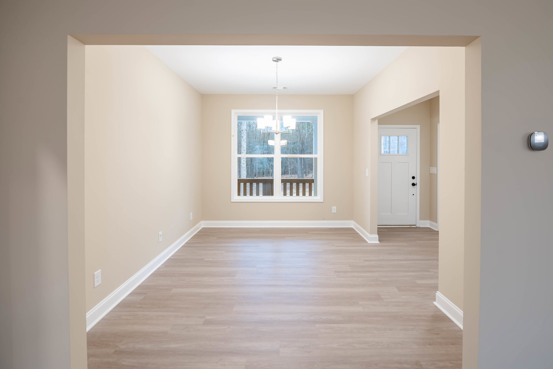 Hardwood floor with white baseboard trim, large window framed by white molding, black hardware on white door, light fixture overhead, trees visible outside