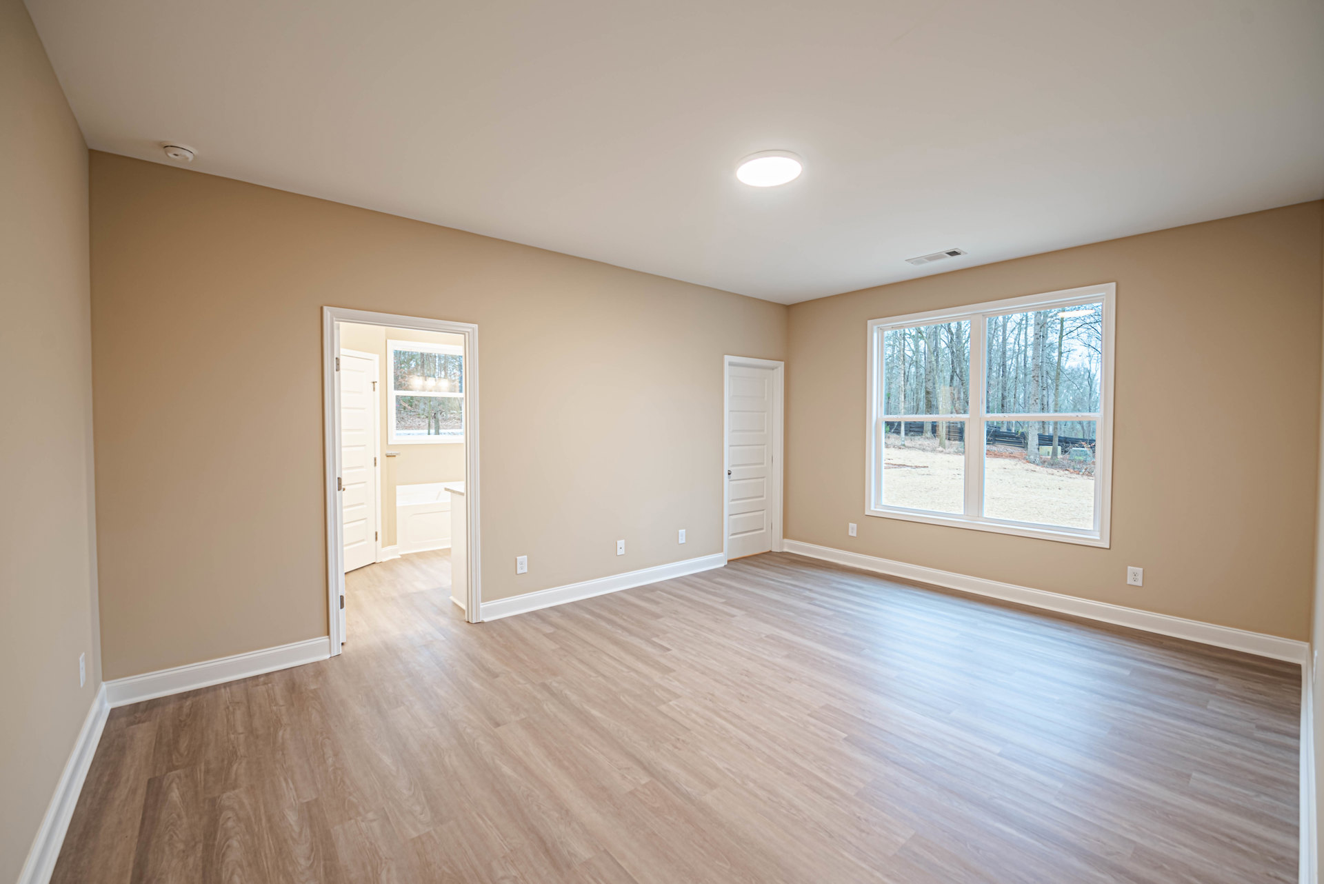 Wood floor room with white walls, ceiling light, large window showing trees outside, and white door with silver handle