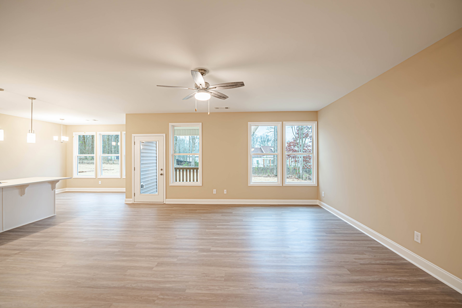 Light wood flooring, white walls with molding, ceiling fan with light fixture, two windows with views of trees and neighboring house, white door with blinds