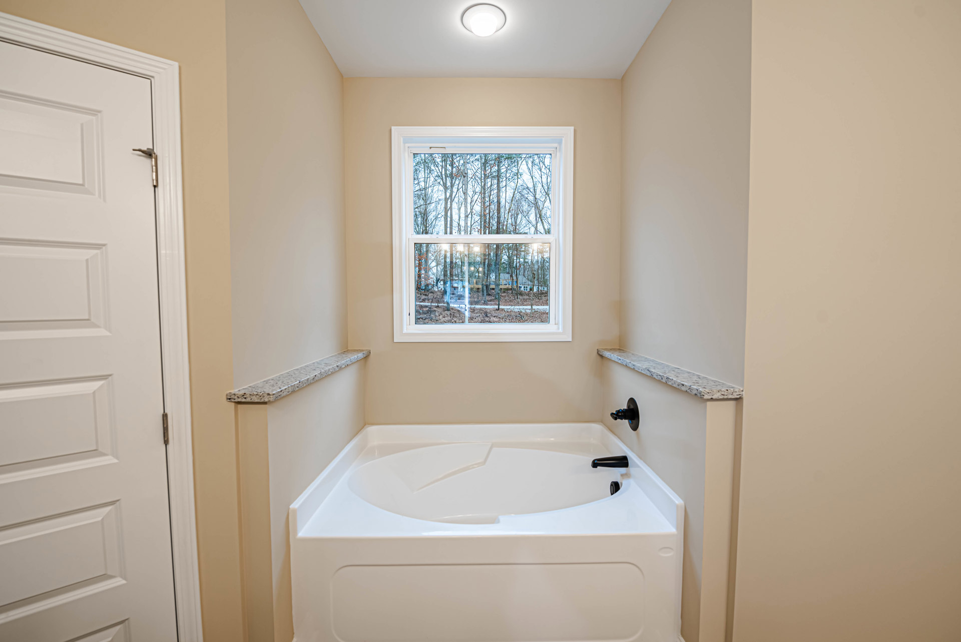 Freestanding white bathtub with matte black faucet, large window overlooking trees, white tile walls, modern light fixture, and white door in contemporary bathroom