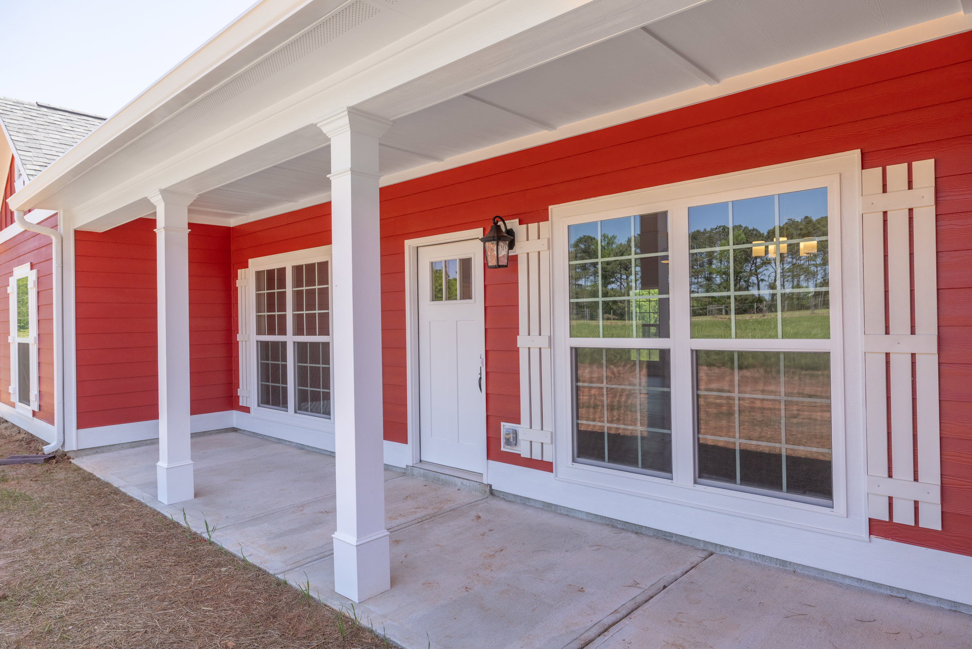 Red siding home with white pillars, white front door, and white-trimmed windows; exterior entryway features a white ceiling and red blinds.