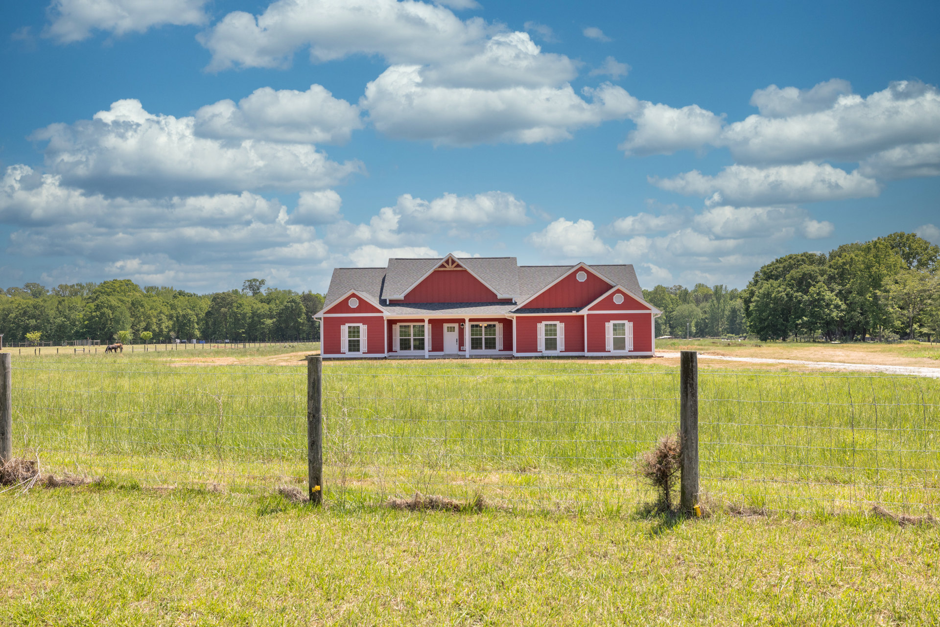 Red house with white trim and red roof set in a grassy fenced field, surrounded by trees under a cloudy sky