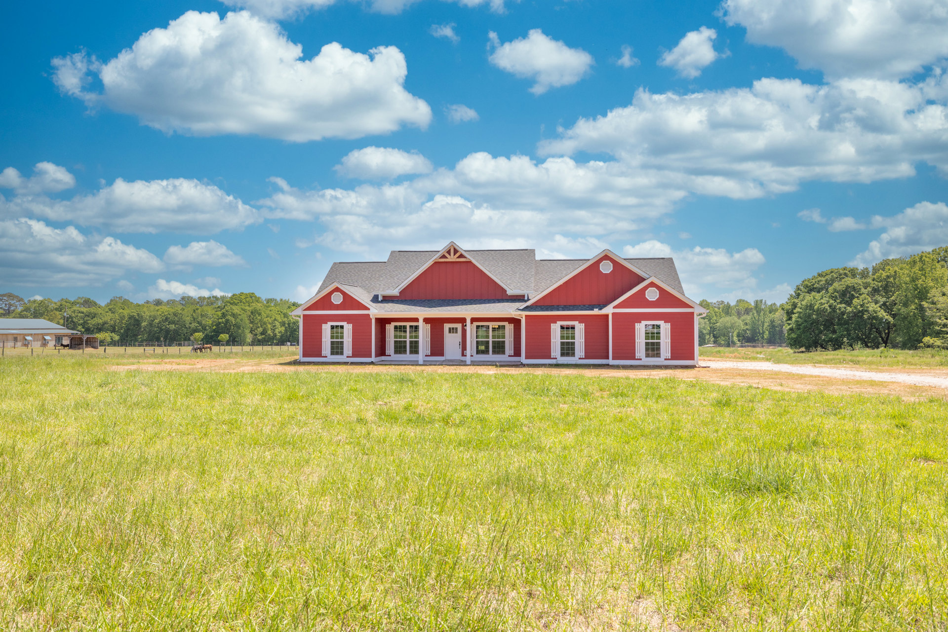Red house with white trim and red roof, surrounded by green grass and open field, white-framed windows, rural landscape with trees and cloudy sky