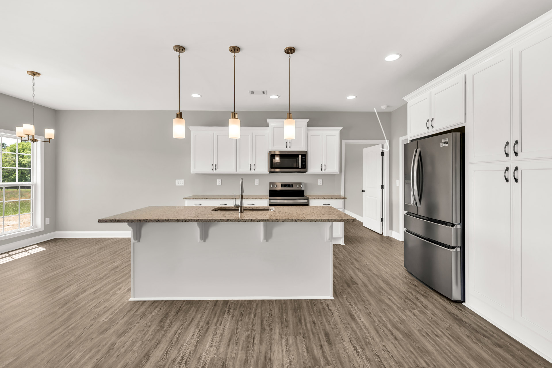 Modern kitchen featuring a central island with white countertops, stainless steel refrigerator, built-in microwave and oven, light cabinetry, sink, and a window overlooking trees.