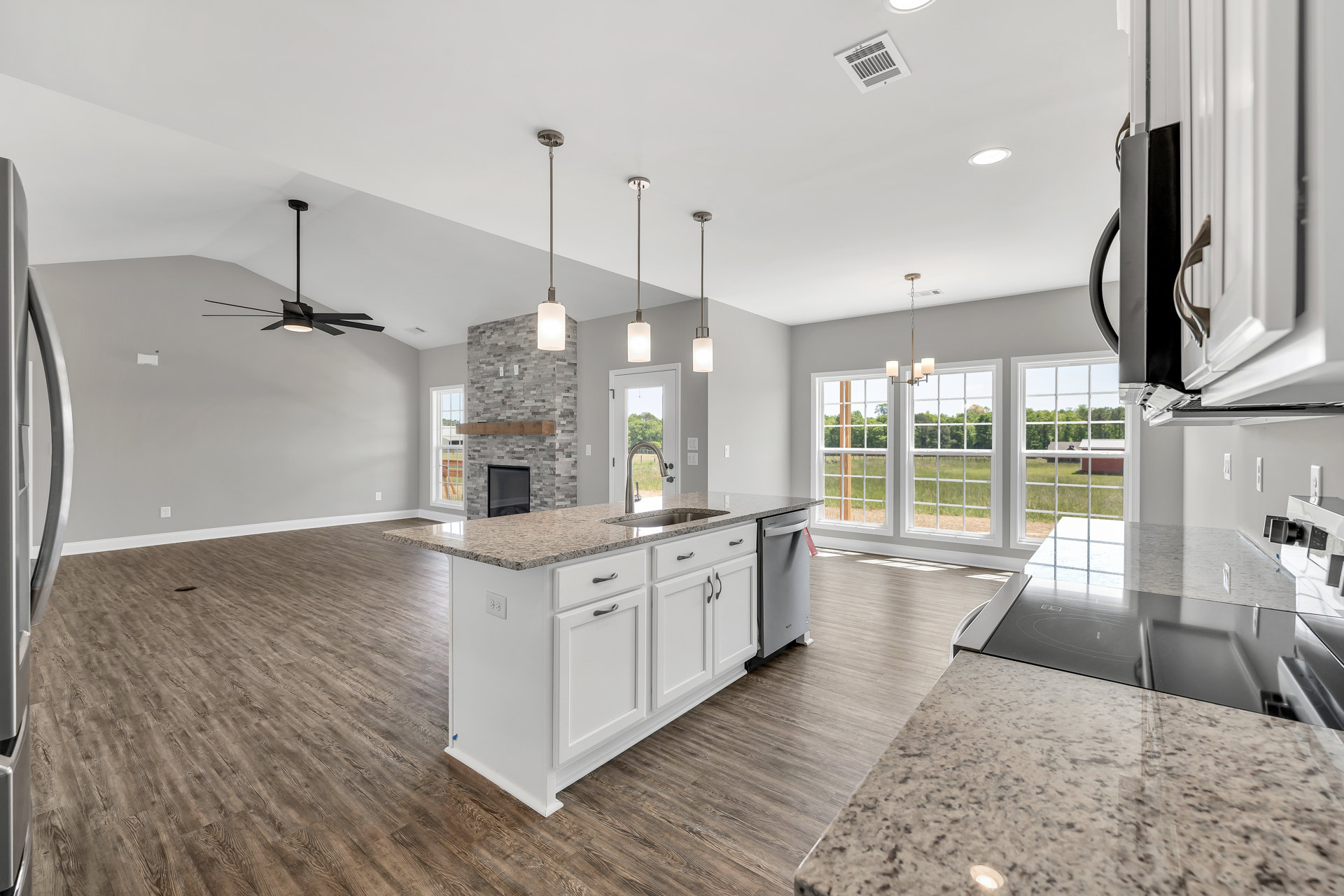 Spacious kitchen featuring white cabinets, granite countertops, large central island, tile flooring, ceiling fan with light, glass-door fireplace, stainless steel sink, and modern
