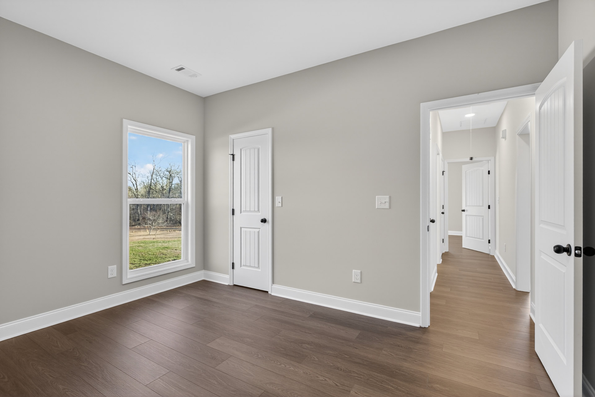 White paneled door with black knobs, wood laminate flooring, white walls, large window showing trees outside, grey countertop along one wall.