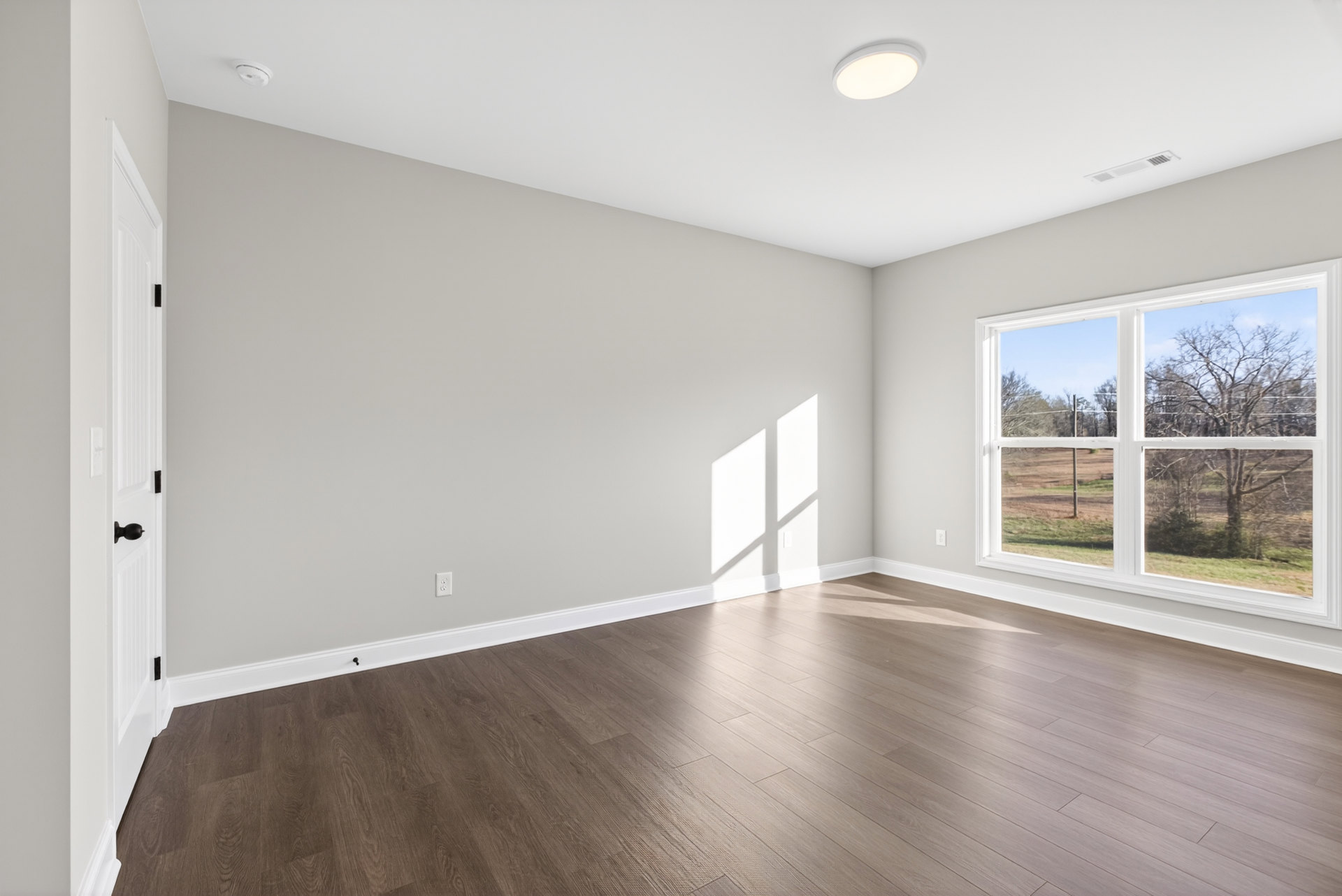 Sunlit room with wide wood plank flooring, large window overlooking leafy trees, white walls and ceiling, recessed lighting, and a light switch near the doorway