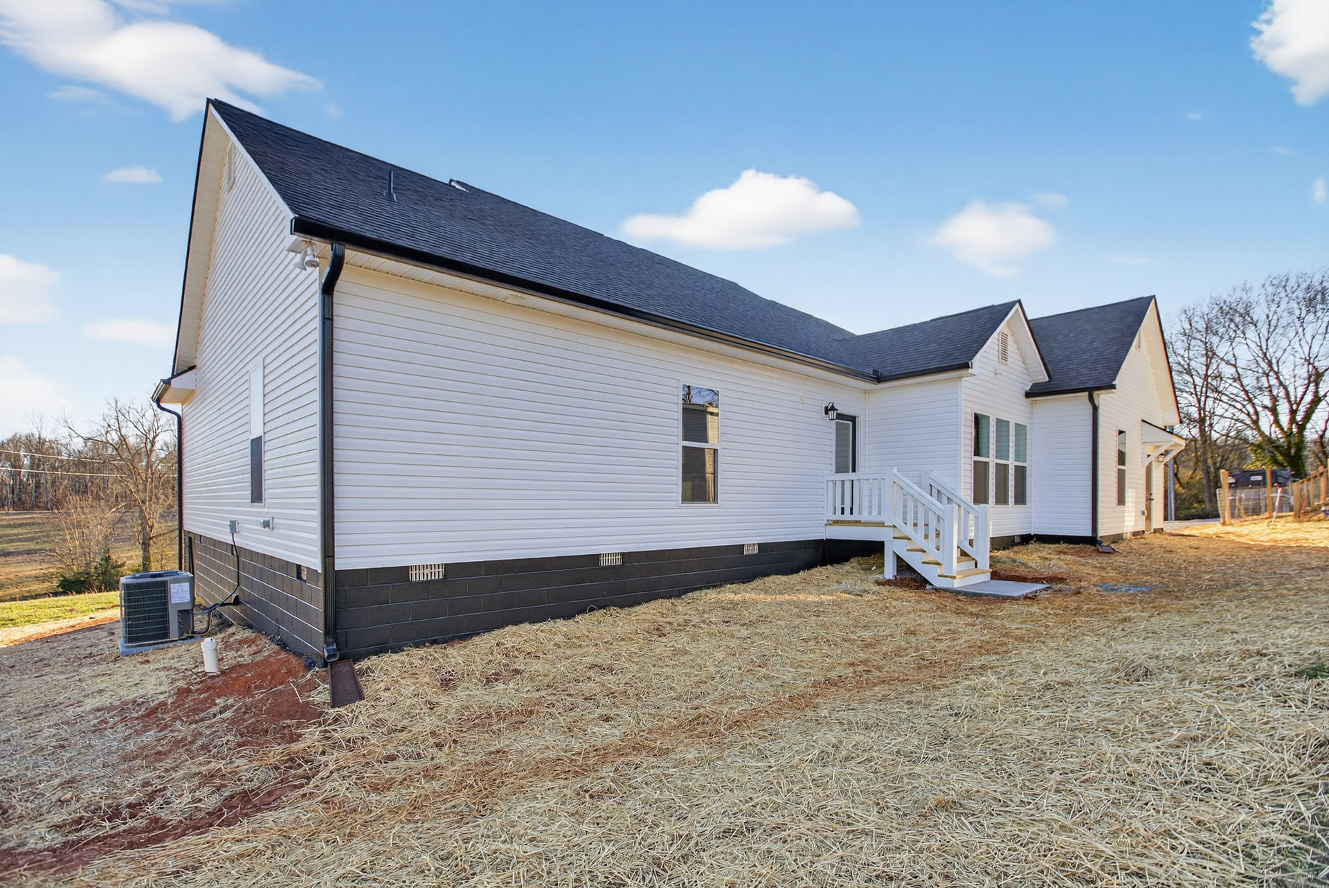 White siding house with black shingle roof, white framed windows, white exterior staircase, hay scattered on ground, large grey HVAC unit with wire mesh, surrounded by trees under