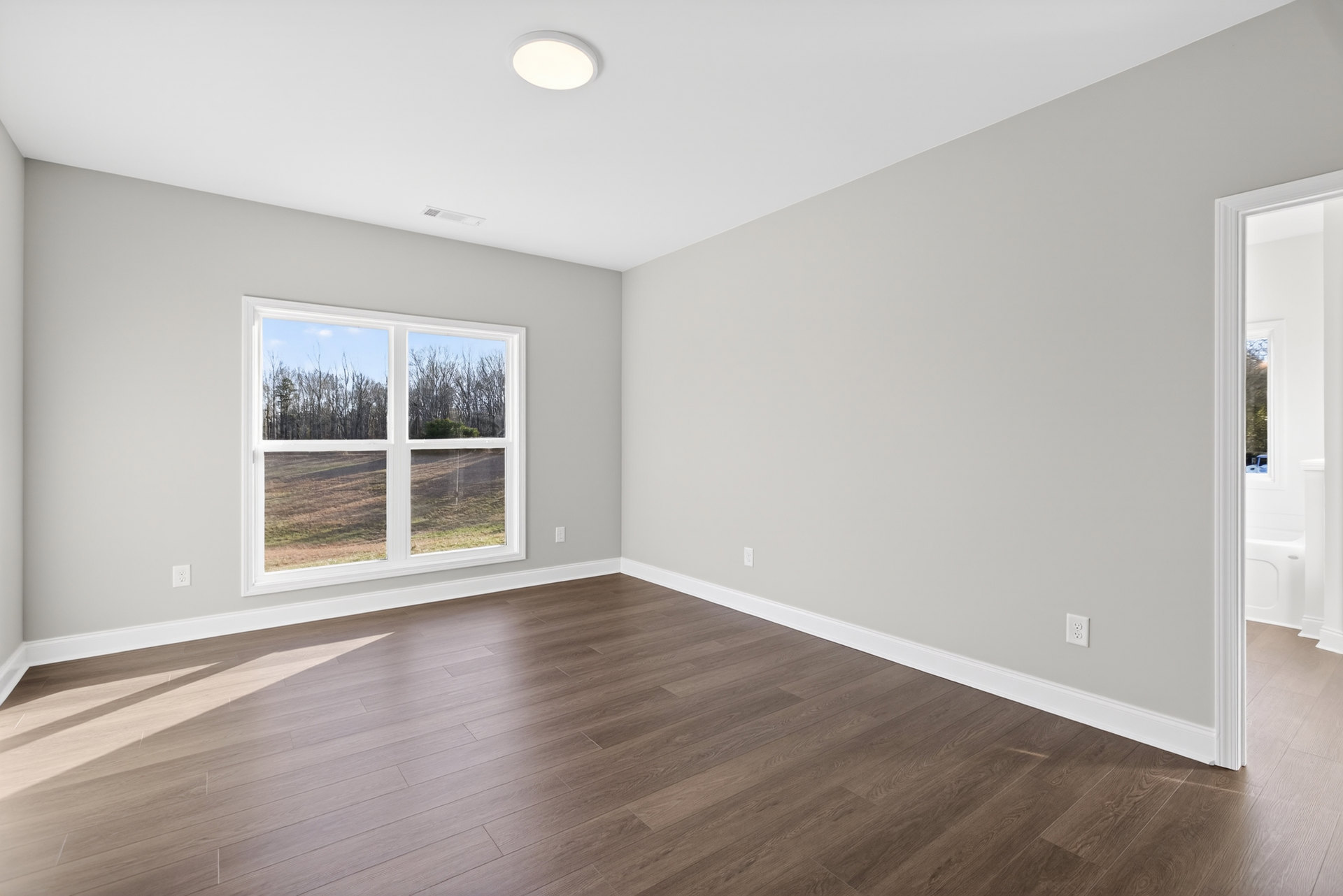 Sunlit room with wide window overlooking leafy trees, smooth wood flooring with light reflections, white plaster walls, and minimalist interior design.