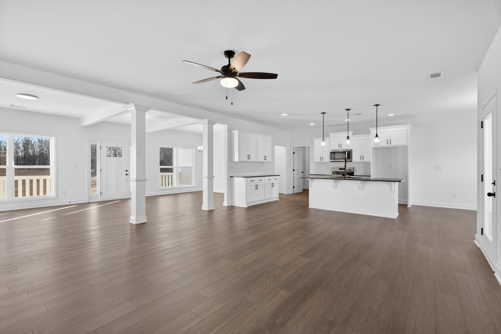 Spacious white kitchen and living area with wood flooring, ceiling fan with light, white cabinets, black countertops, large window overlooking trees, and white kitchen counter with