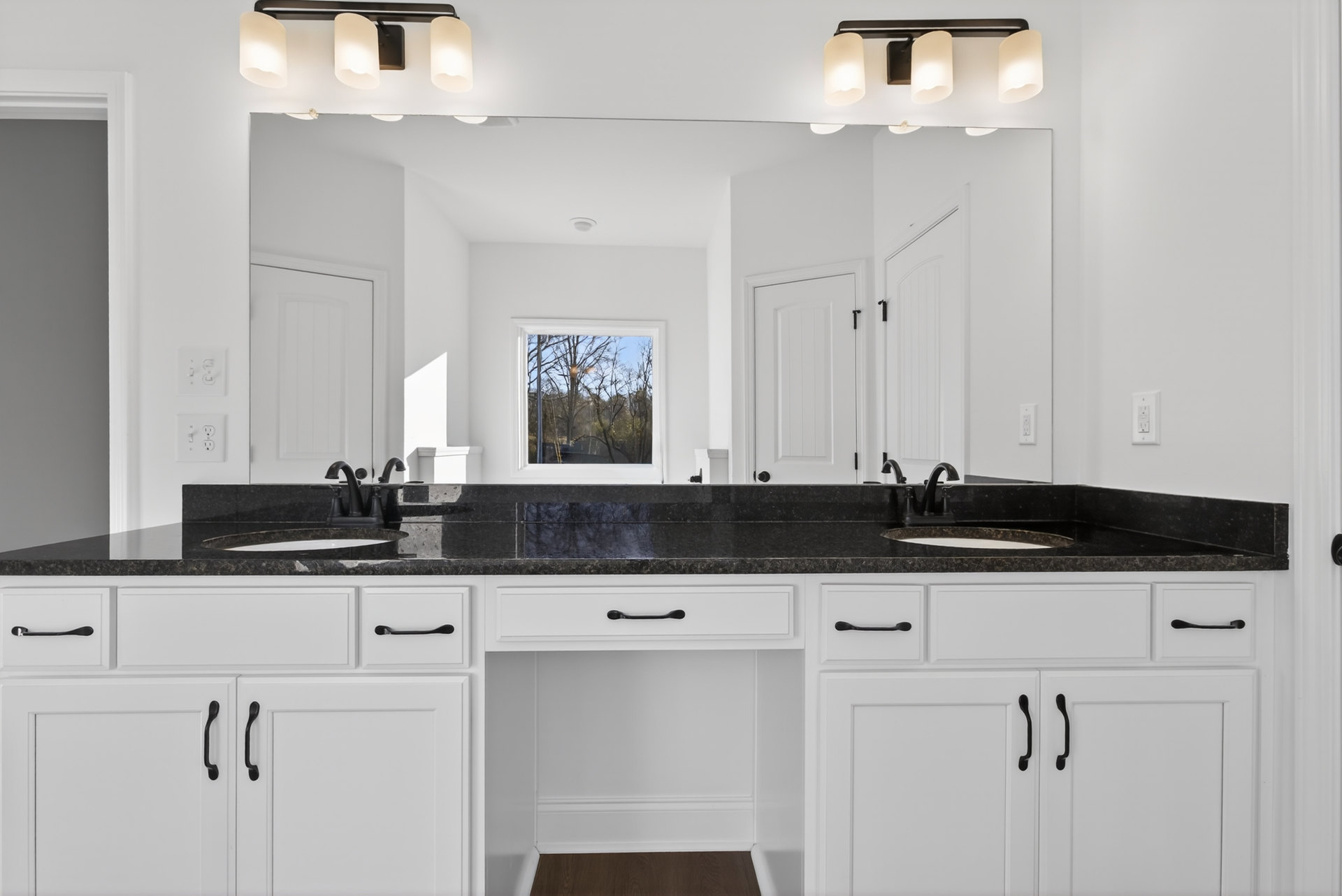 Bathroom with expansive mirror above white countertop and sink, black hardware on white cabinetry, window framing leafy trees, wood accent surface, and paper on counter