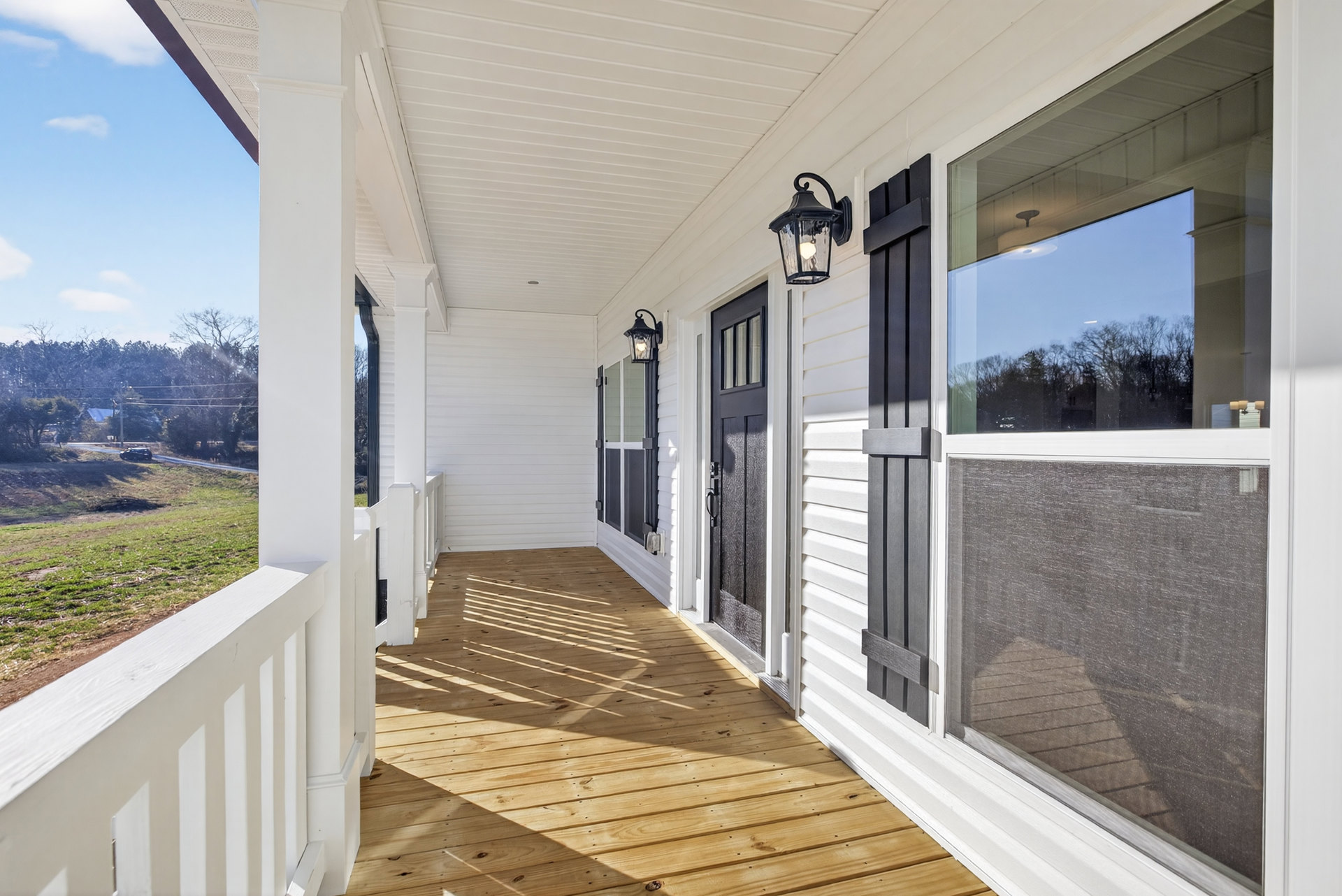White porch with wood decking, white railing, black front door, window reflecting trees, and outdoor light fixture; grass and dirt visible beyond railing.