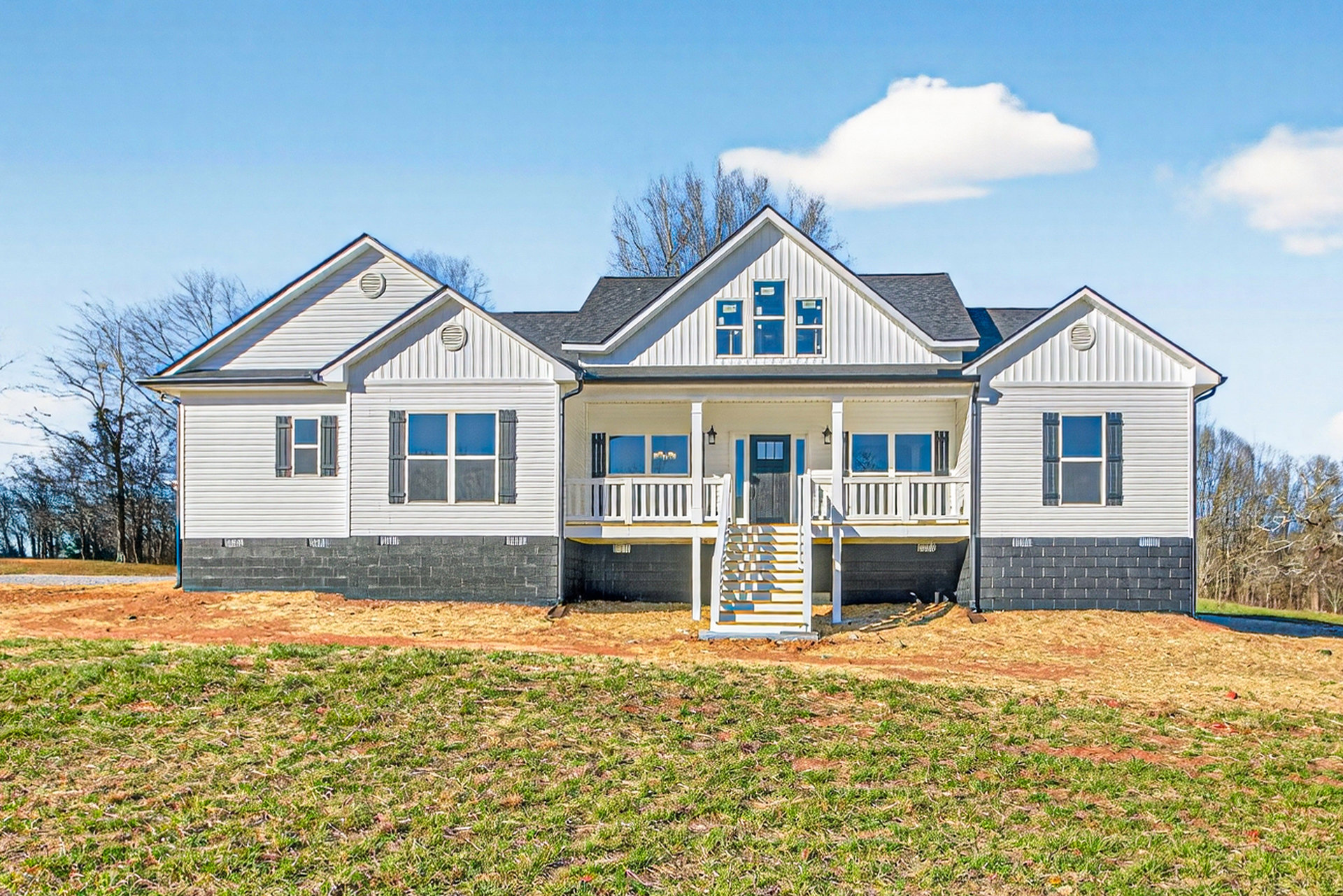 White two-story house with blue shutters, wide front porch, and white staircase, surrounded by a large green lawn and mature trees under a partly cloudy sky