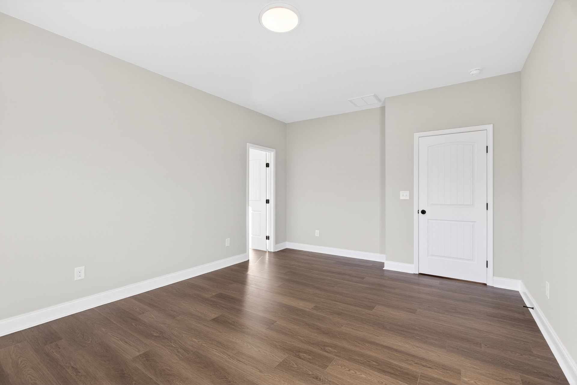 White paneled door with black knob opening onto a room with light wood laminate flooring, smooth white plaster walls, and a ceiling-mounted light fixture.