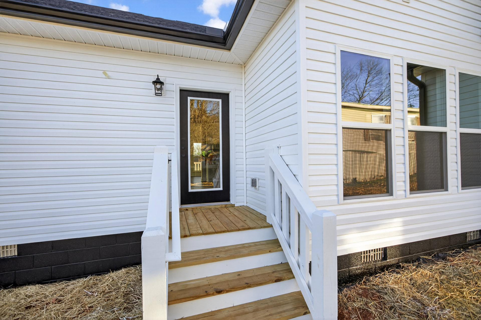 White siding exterior with wooden front door and adjacent glass door, black door and wooden stairs, large windows reflecting tree branches, porch entry, curtain visible through