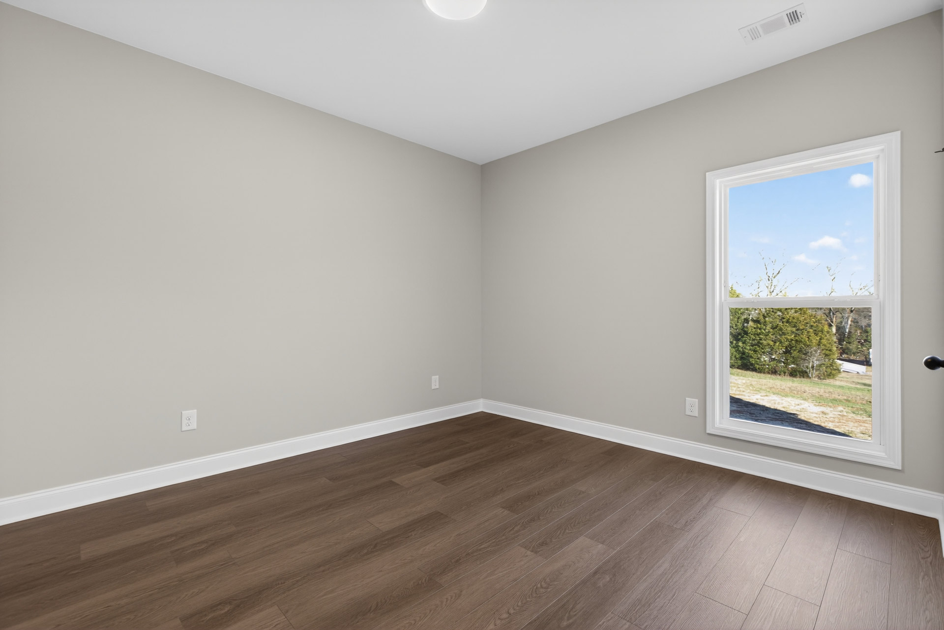 Sunlit room featuring a large window overlooking leafy green trees, hardwood flooring with white baseboard trim, and a rectangular floor vent near the wall.