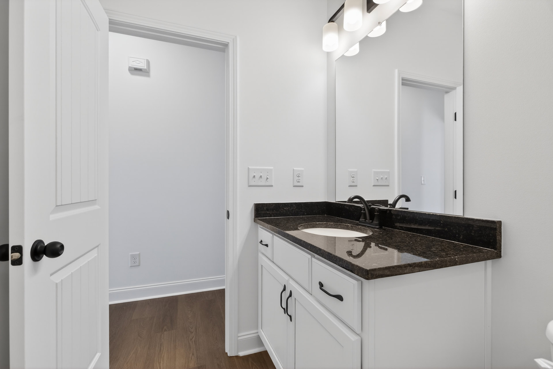 Bathroom with black countertop, undermount sink, chrome faucet, white cabinetry, wood flooring with white trim, and a white door featuring a black handle.