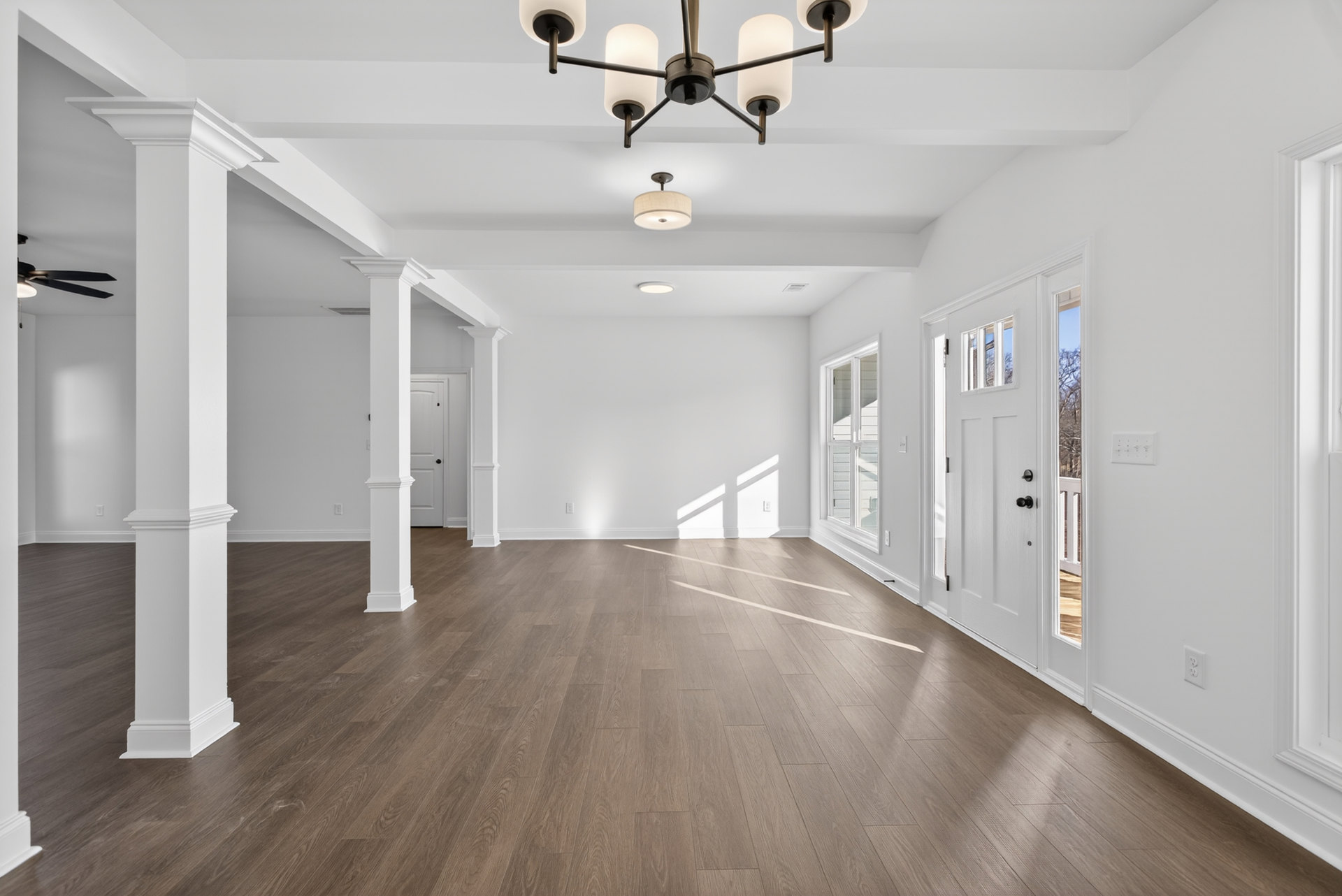 White walls and wood flooring in a bright room with a ceiling fan and chandelier, featuring large windows and a modern light fixture.