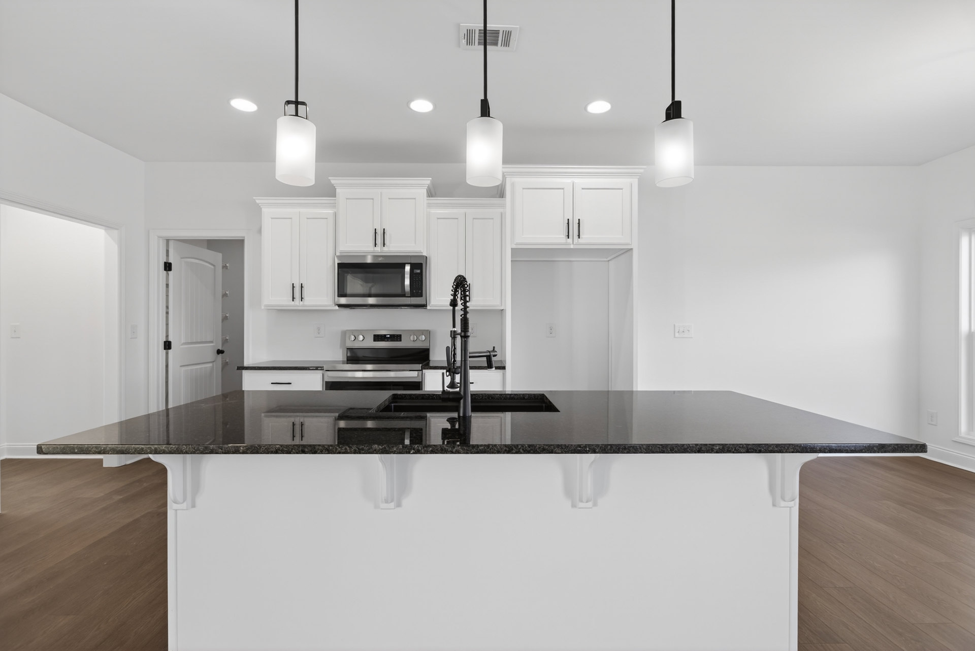 Modern kitchen featuring black countertop, black sink on central island, stainless steel microwave and oven, white cabinetry with black handles, pendant lamp overhead, light wood