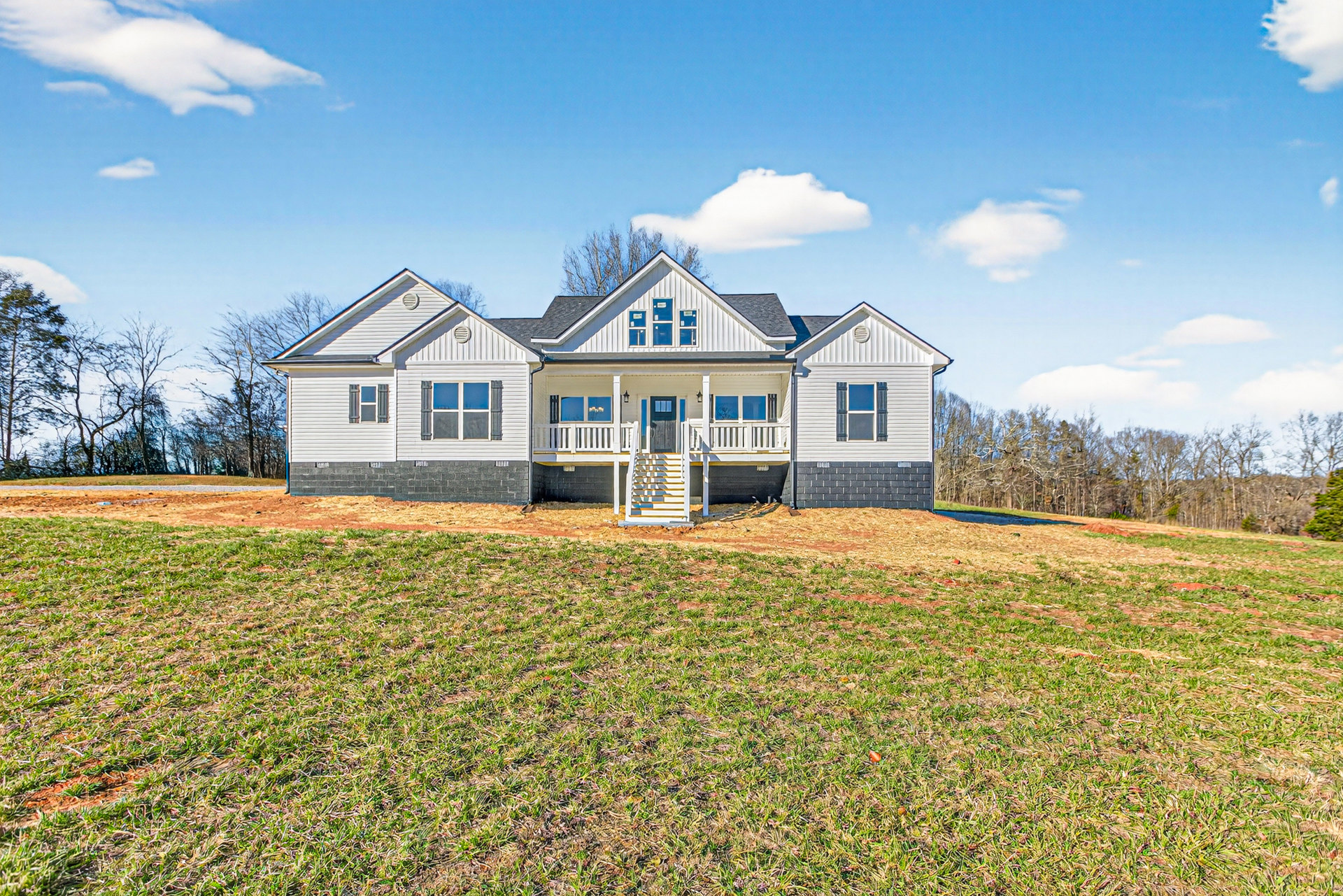 White farmhouse with blue sky, large green lawn, front porch with white stairs featuring yellow and blue stripes, small outbuilding in the grass, scattered trees, and a single