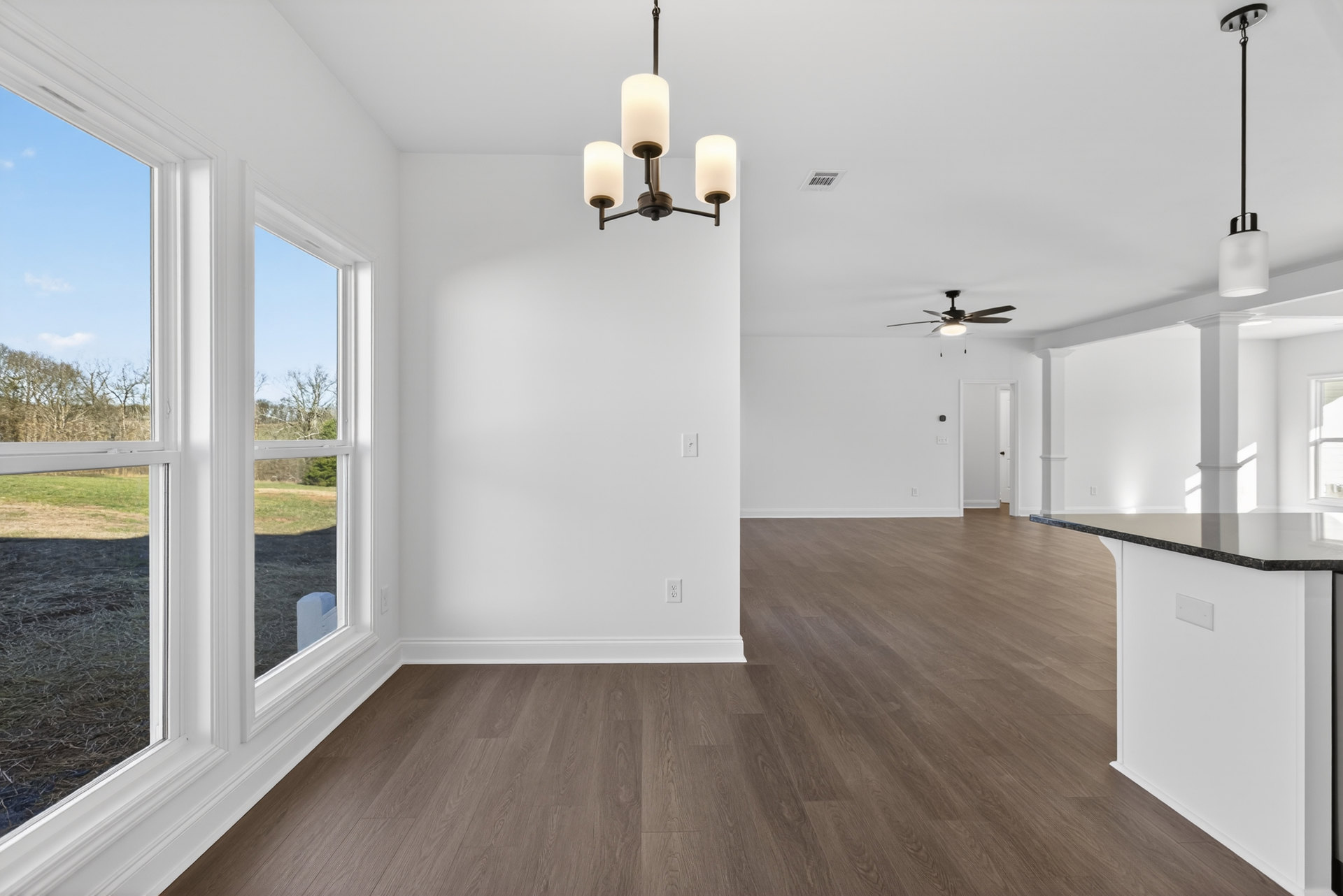 Spacious room with warm wood flooring, white walls, large window, ceiling fan, and chandelier.