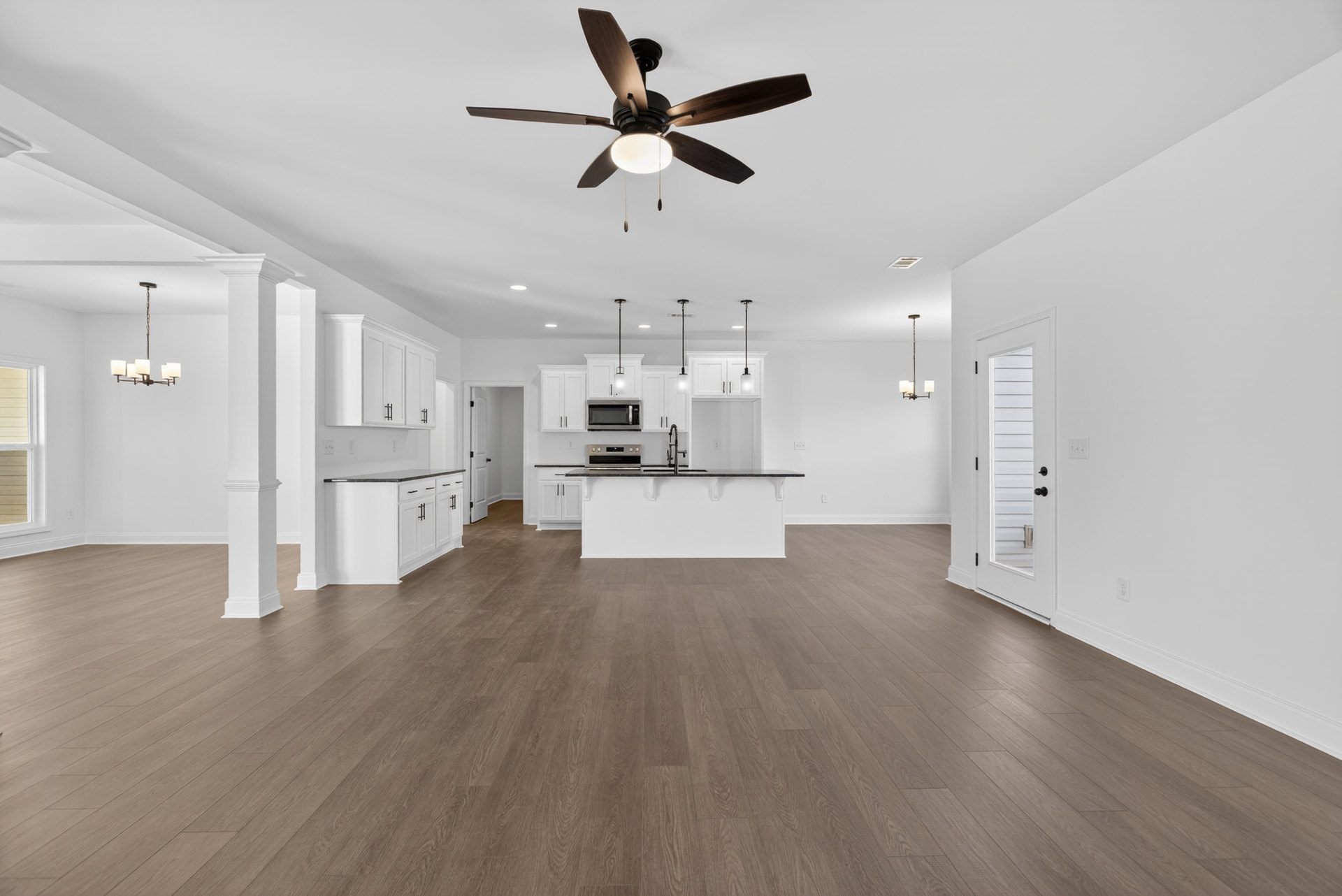 Open kitchen with white cabinetry, black countertops, hardwood flooring, ceiling fan with light, white door with black hardware, and modern chandelier.