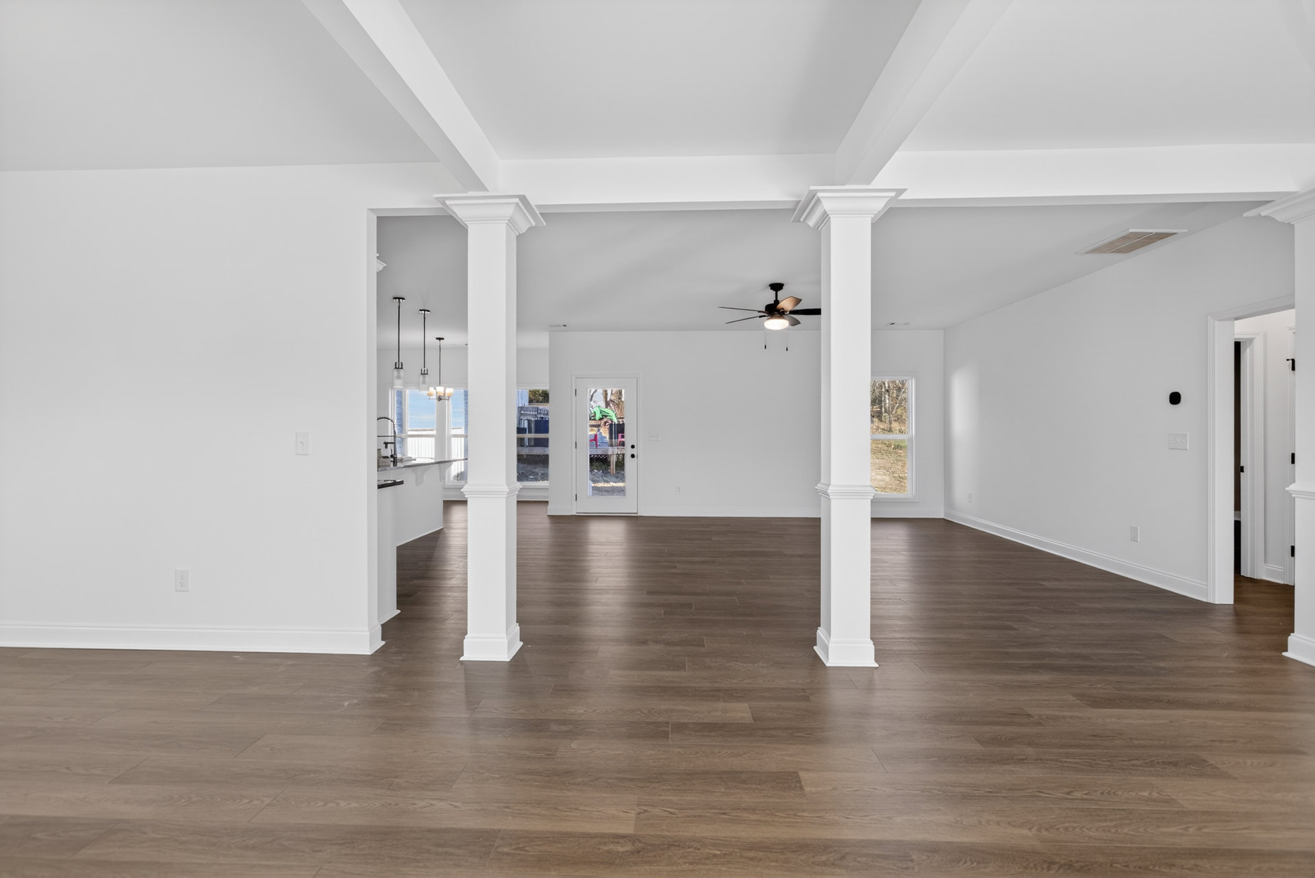 Wood flooring and white pillars in a spacious room, white walls with windows, ceiling fan with light fixture, white door with glass panel, partial view of a fence