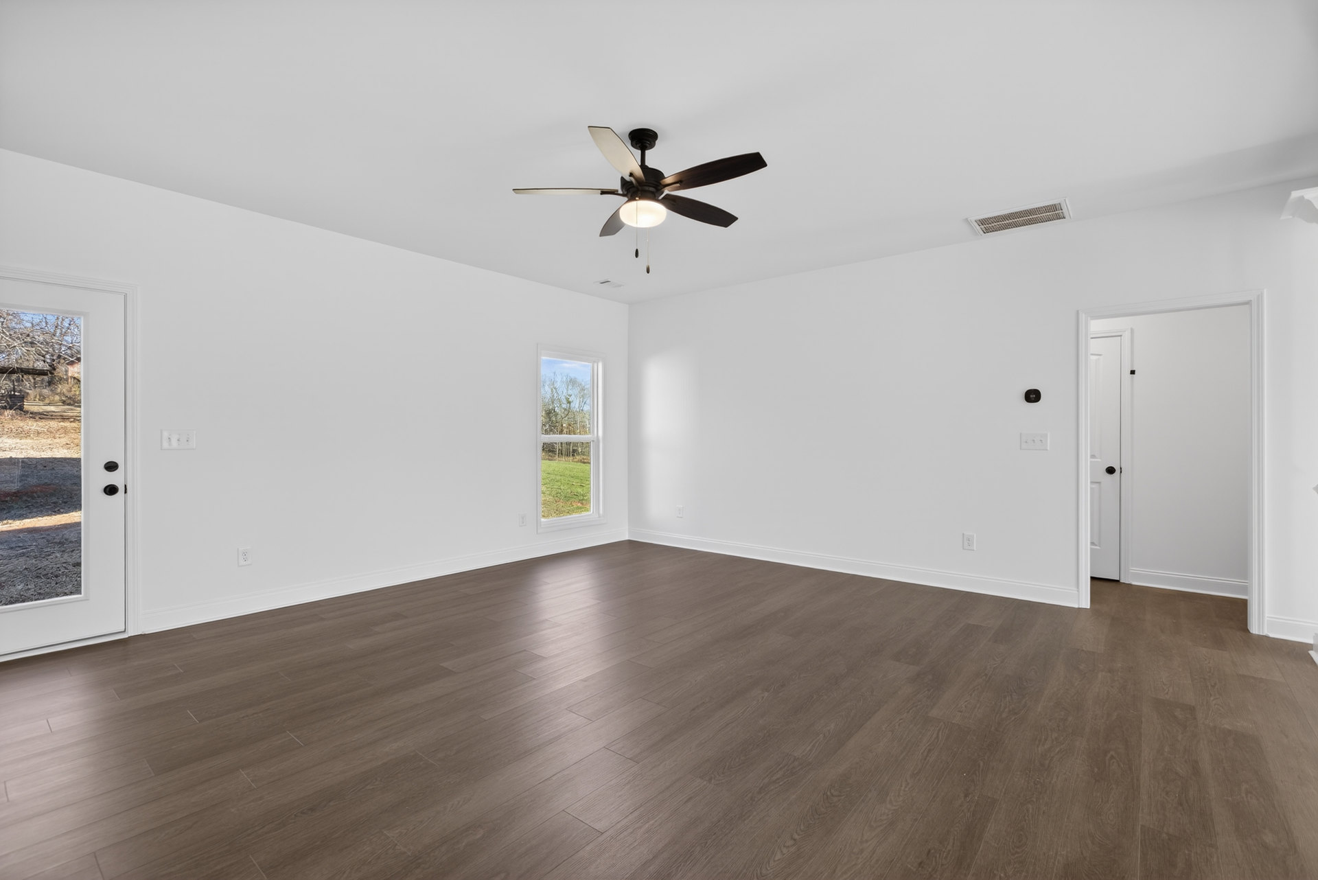 Ceiling fan with light fixture above hardwood floors, white plaster walls, large window overlooking grassy field, floor vent near baseboard