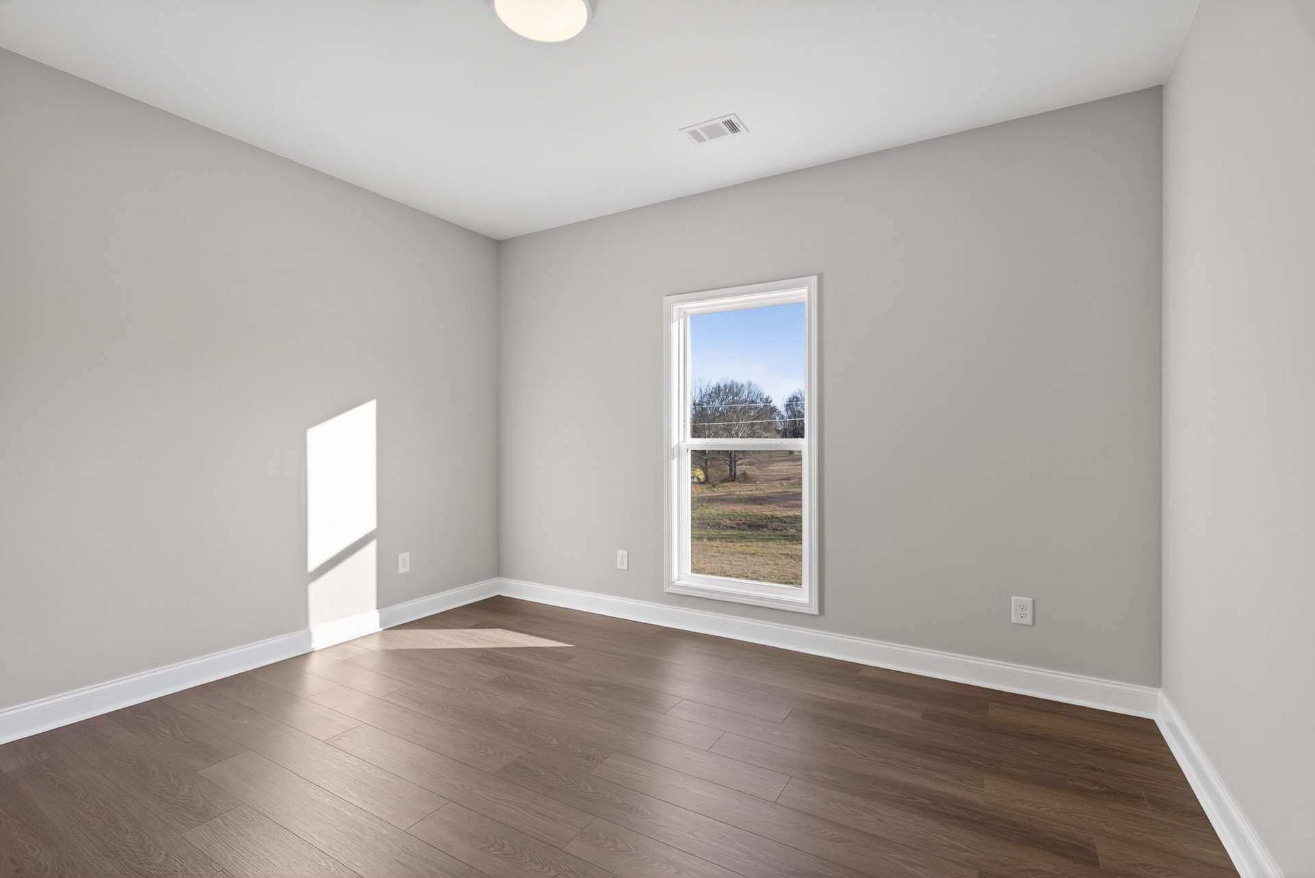 Sunlit room with wide window overlooking field and trees, white plaster walls, rich hardwood flooring, and window shadow cast across floor and wall