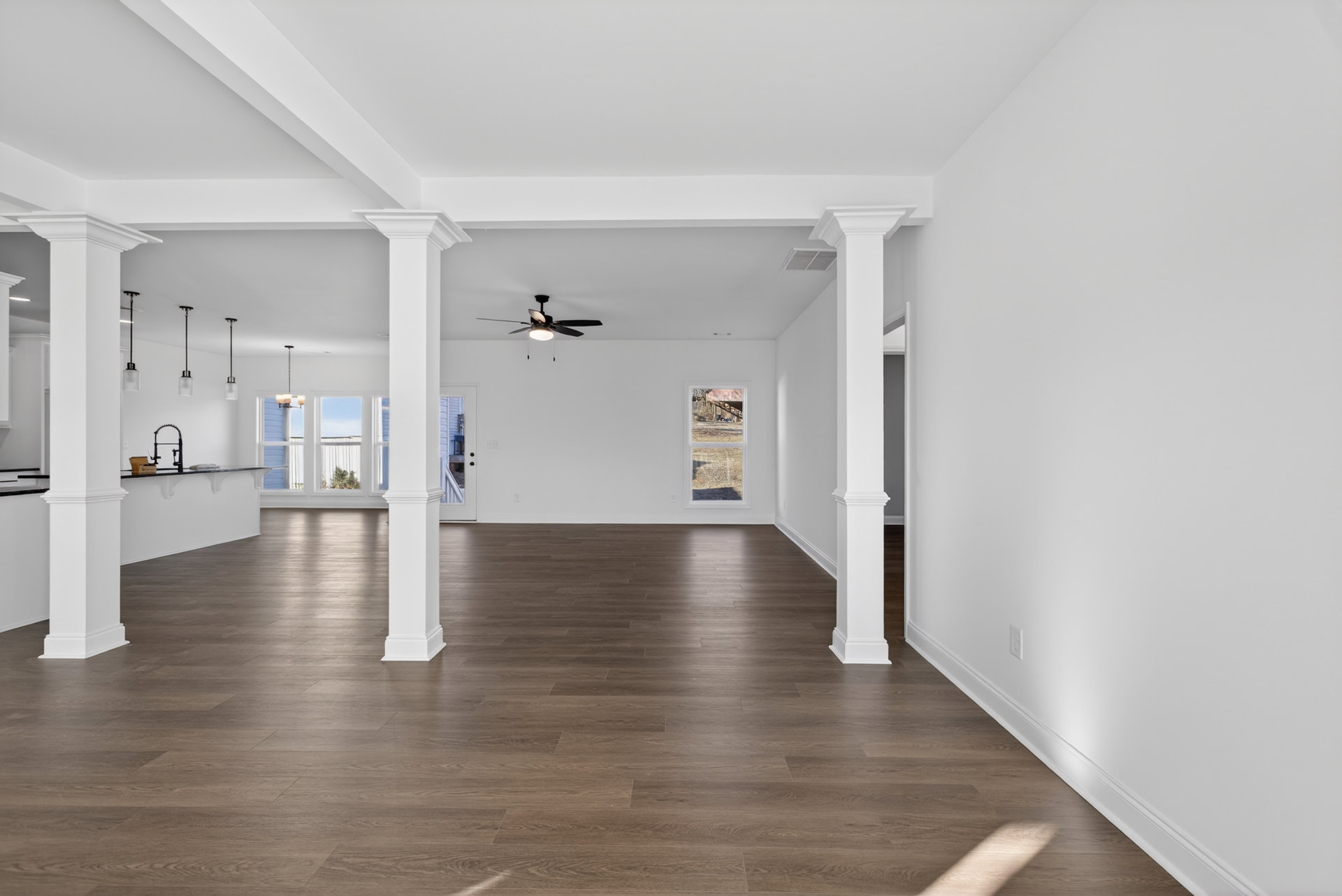 Spacious room with hardwood floors, white pillars, ceiling fan with light, white walls, black door, and window overlooking a grassy field