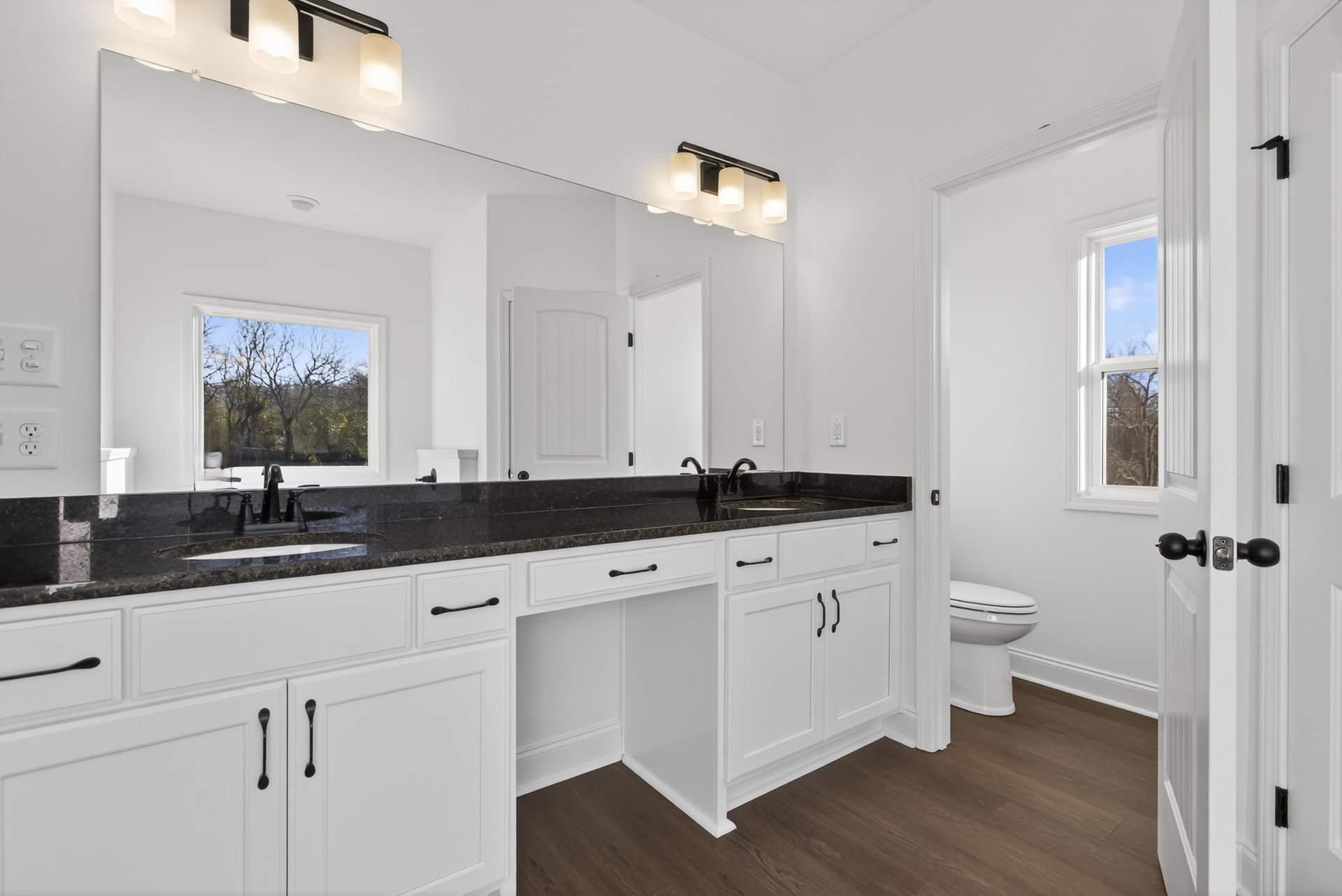 Bathroom with white cabinets, black stone countertop, undermount sink, chrome faucet, toilet with closed lid, window showing trees, row of bulb light fixtures above mirror, tile