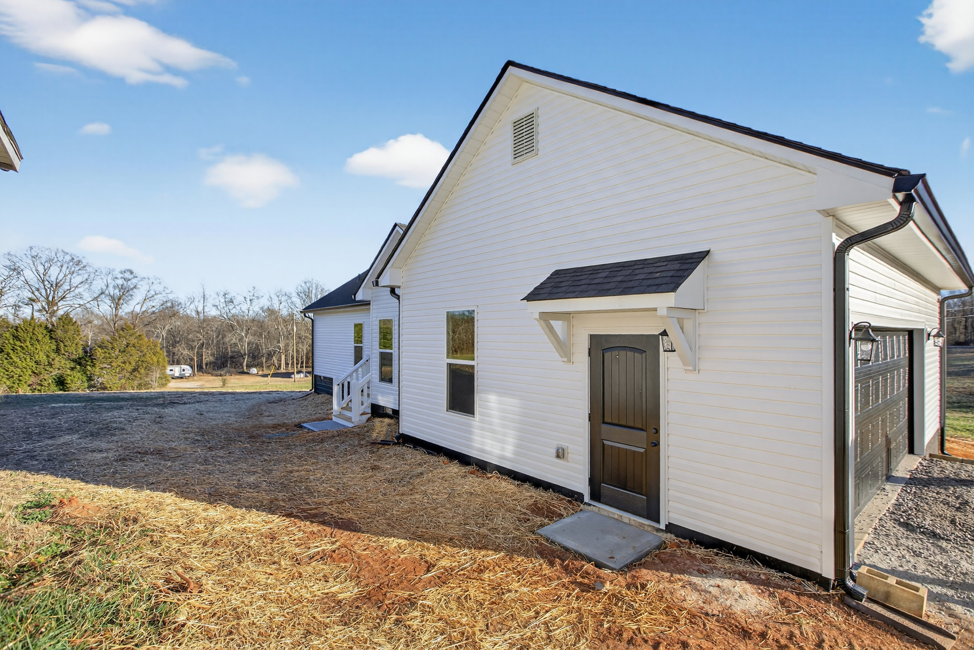 White siding house with brown front door, gravel yard, and large windows; cloudy sky and trees in background