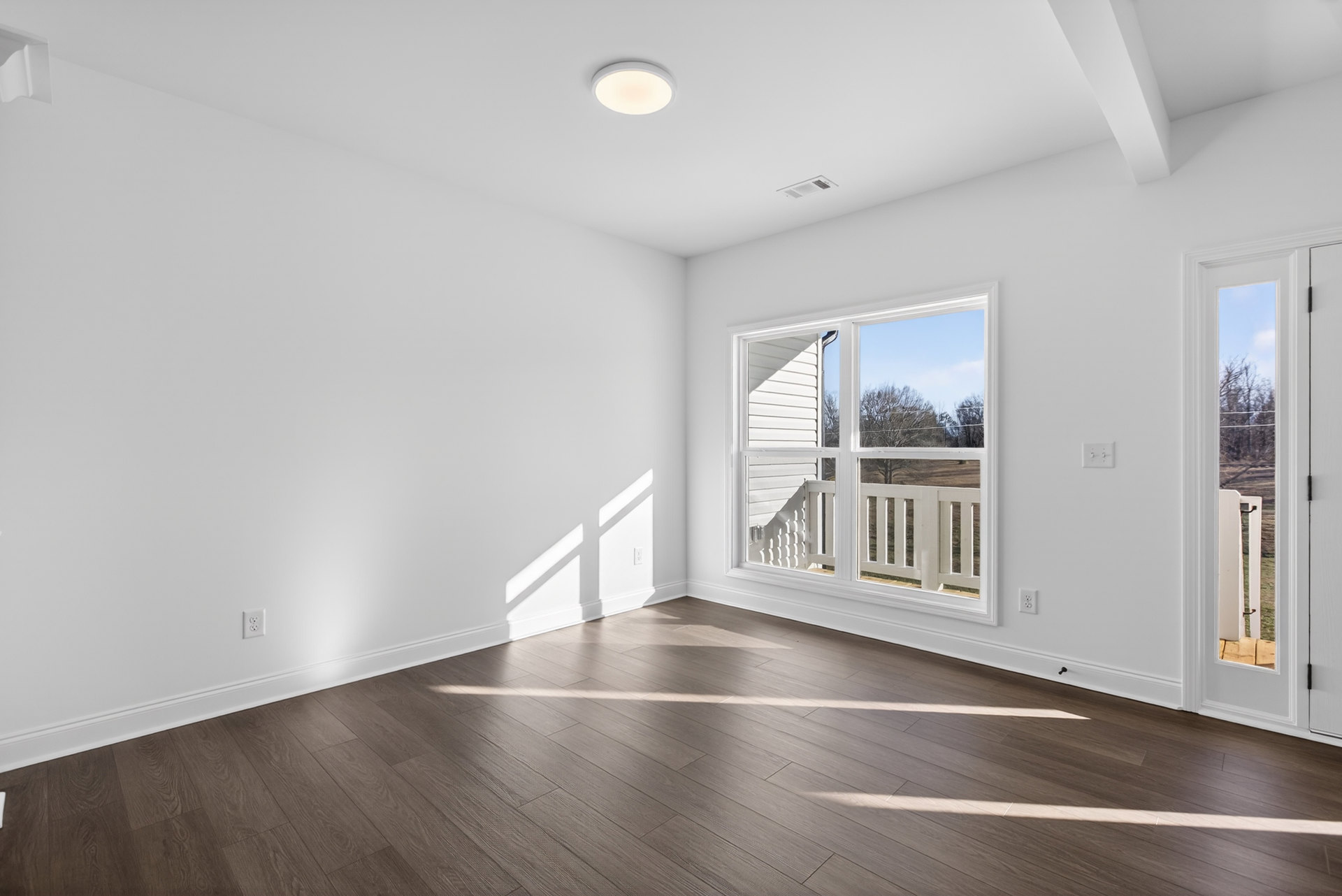 Sunlit room with wide wood plank flooring, white plaster walls, large window overlooking grassy field, and white railing visible outside