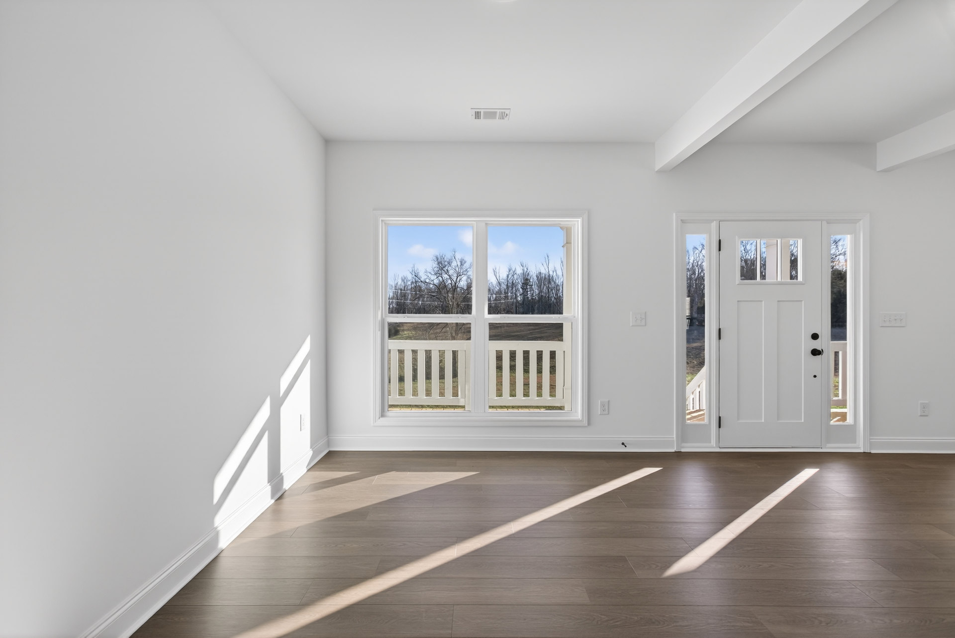 Sunlit room featuring wide plank wood flooring, large window with tree and sky view, white walls with plaster molding, and a white door with glass panes.