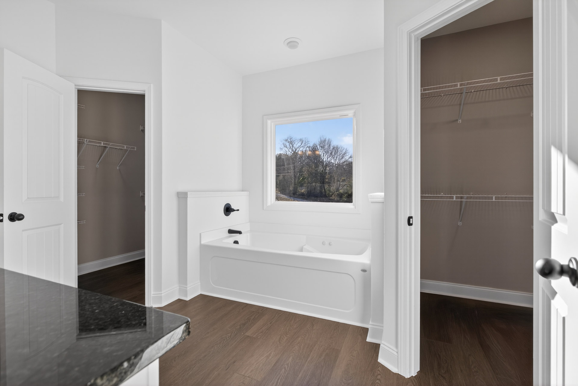 Bathroom with a freestanding white bathtub featuring a black faucet, wood flooring, large window overlooking trees, and a black countertop visible in the adjacent kitchen area.