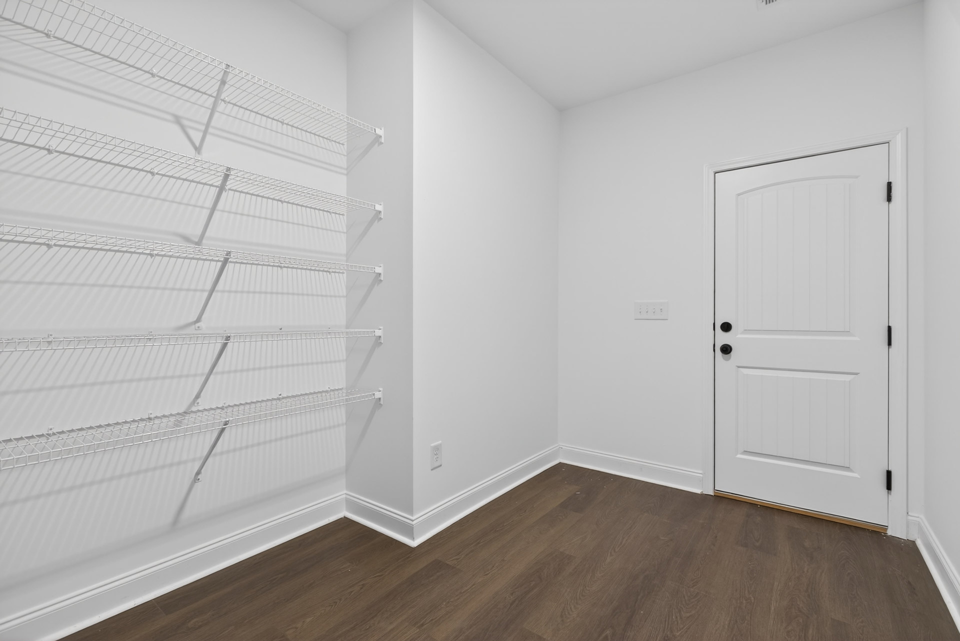 White paneled door with black knobs, built-in white shelves on wall, light wood flooring, smooth white plaster walls and ceiling.