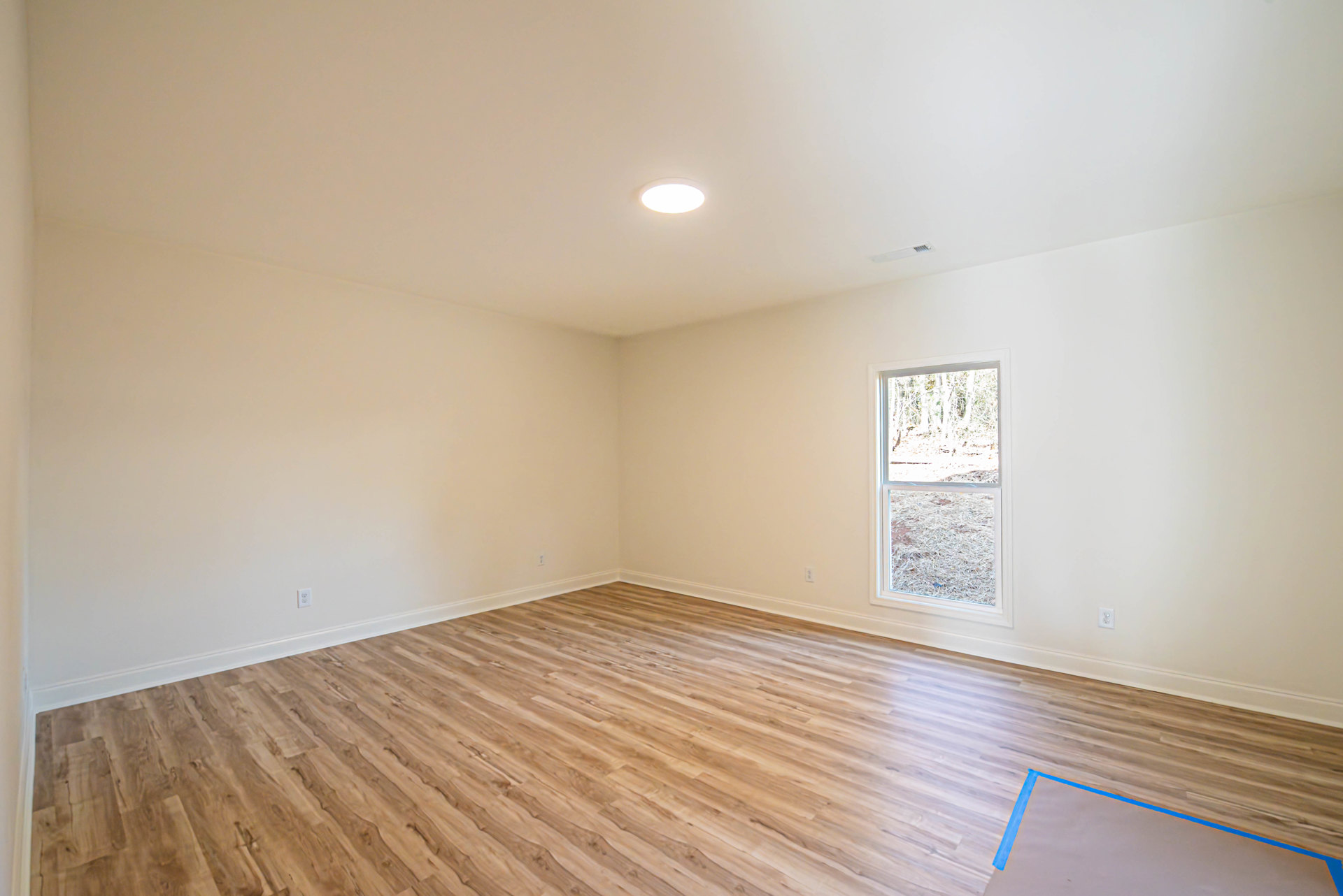 Wood flooring in a room with white walls, ceiling light fixture, and a large window framed in white overlooking a forest.