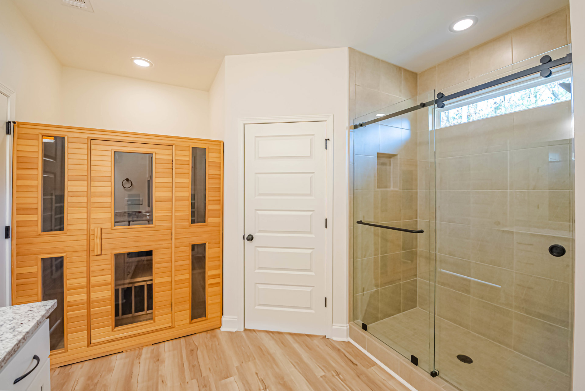 Bathroom with glass shower enclosure, wood vanity cabinet, white door with black knob, tiled floor and walls, ceiling light fixture, countertop with black handle.
