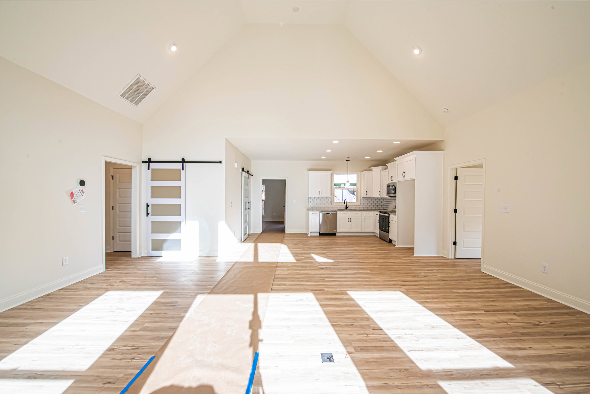 Spacious kitchen with high ceiling, wood flooring, recessed ceiling vent, and overhead lighting; blue painter’s tape visible on floor near white cabinetry