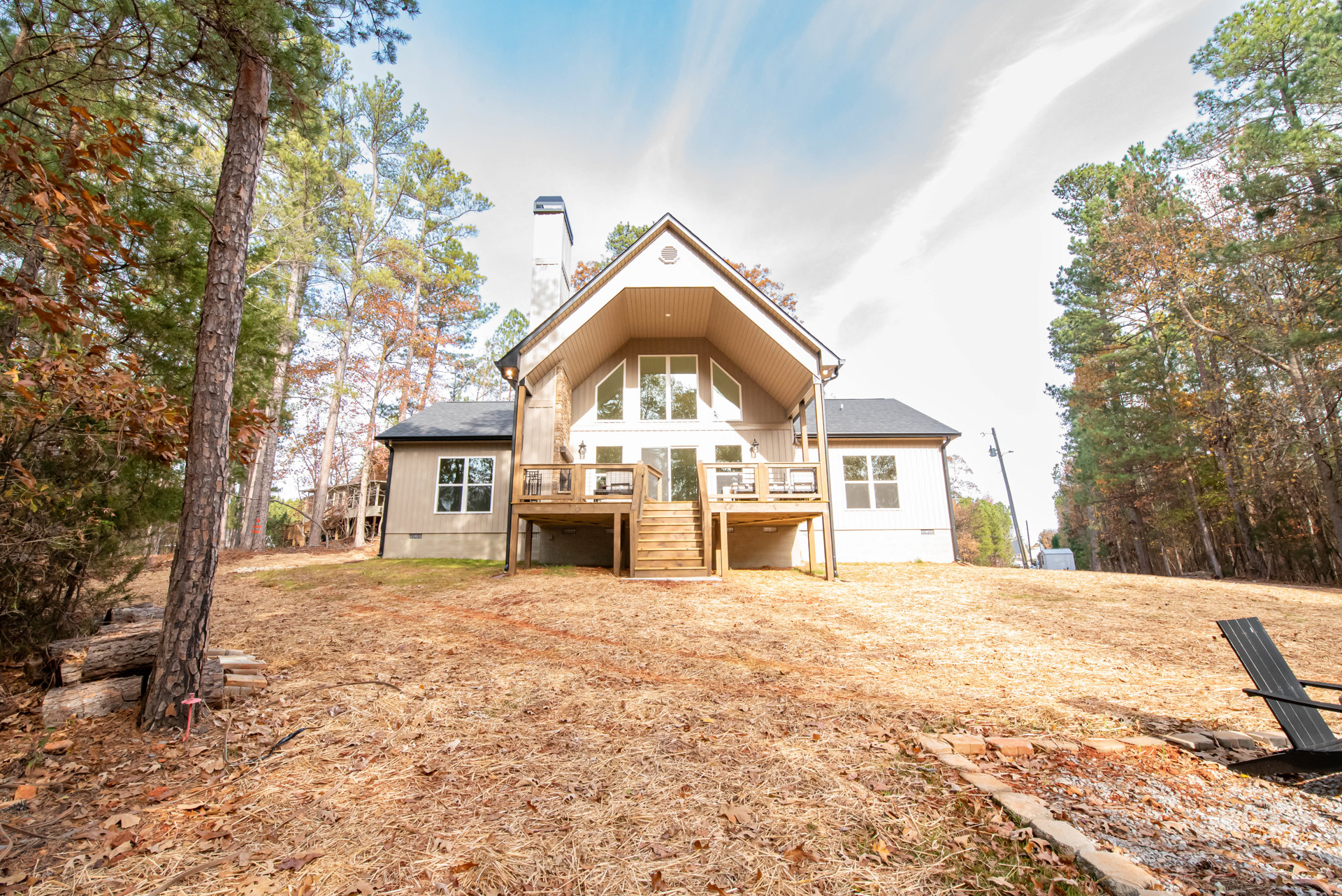 Spacious wooden house with a large covered porch, glass door, wooden stairs, and deck with fence, set in a grassy yard with mature trees and a black outdoor chair