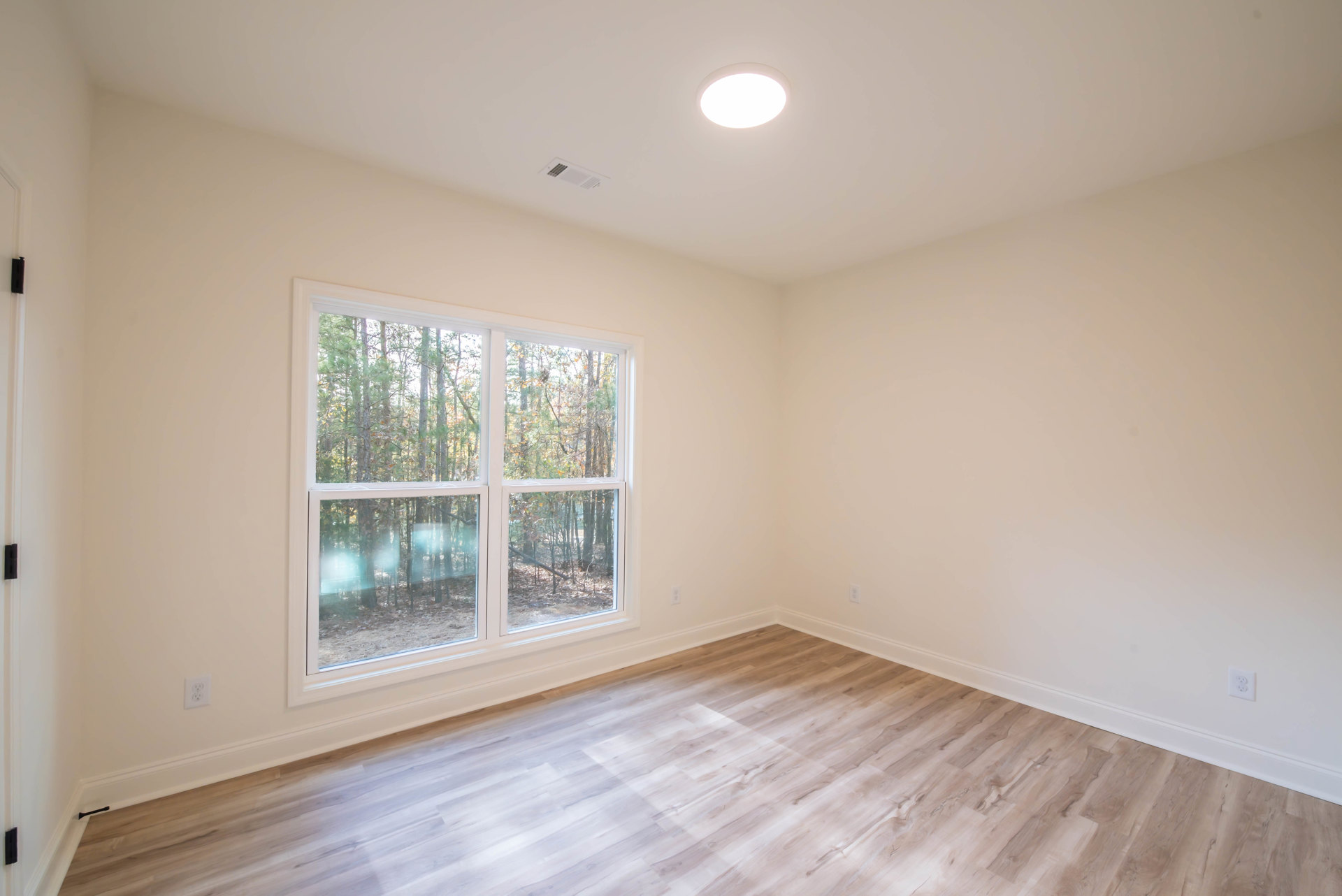 Sunlit room featuring a large window overlooking trees, smooth hardwood flooring, white plaster walls, and recessed ceiling light.